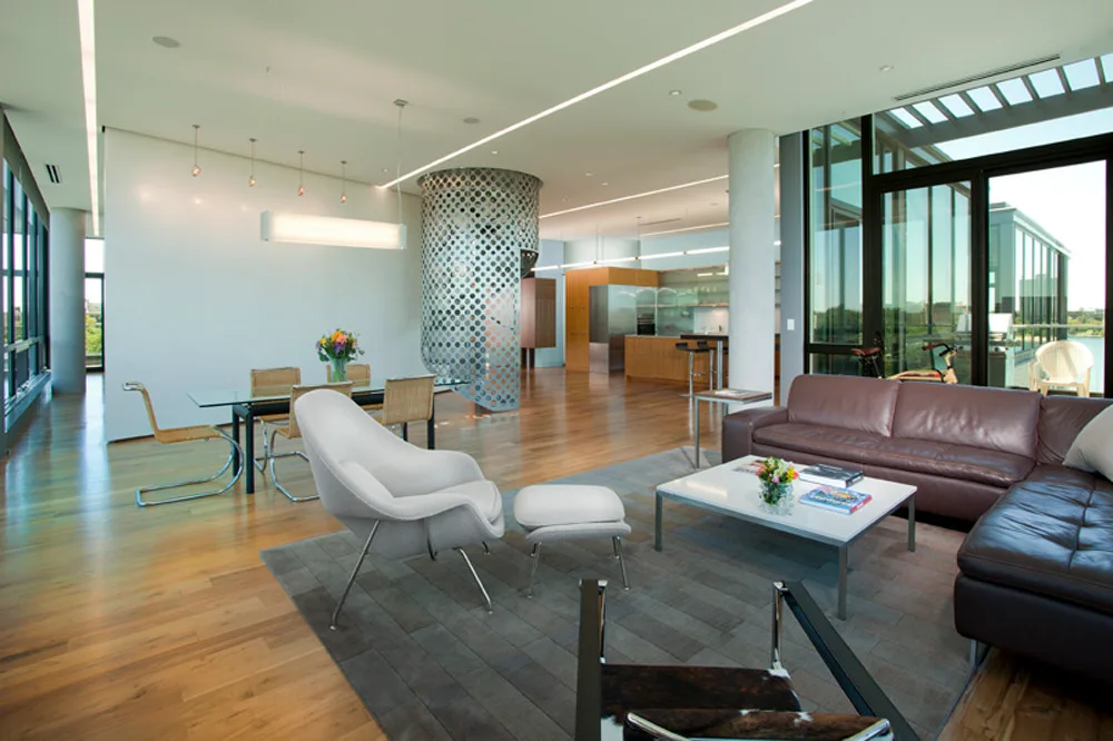 A spacious open-plan living room features a brown leather sectional, a grey lounge chair, and a large perforated metal column near a dining area.