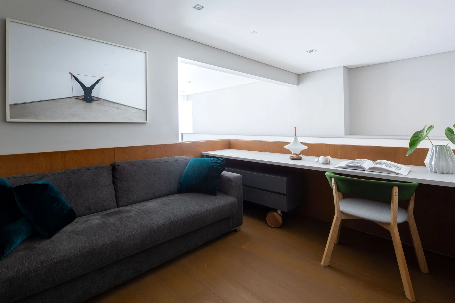A modern home office features a dark grey sofa, a long white desk with a green chair, and wood-toned wall trim.