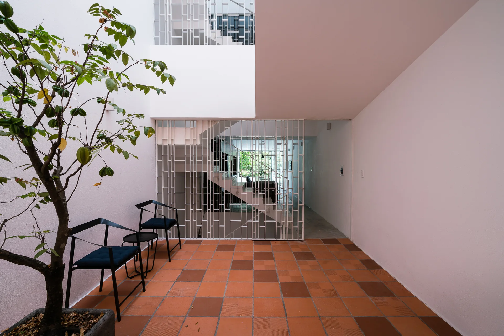 An indoor courtyard with orange terracotta tiles, two black metal chairs, a potted tree, and a white metal grid screen in front of a staircase.