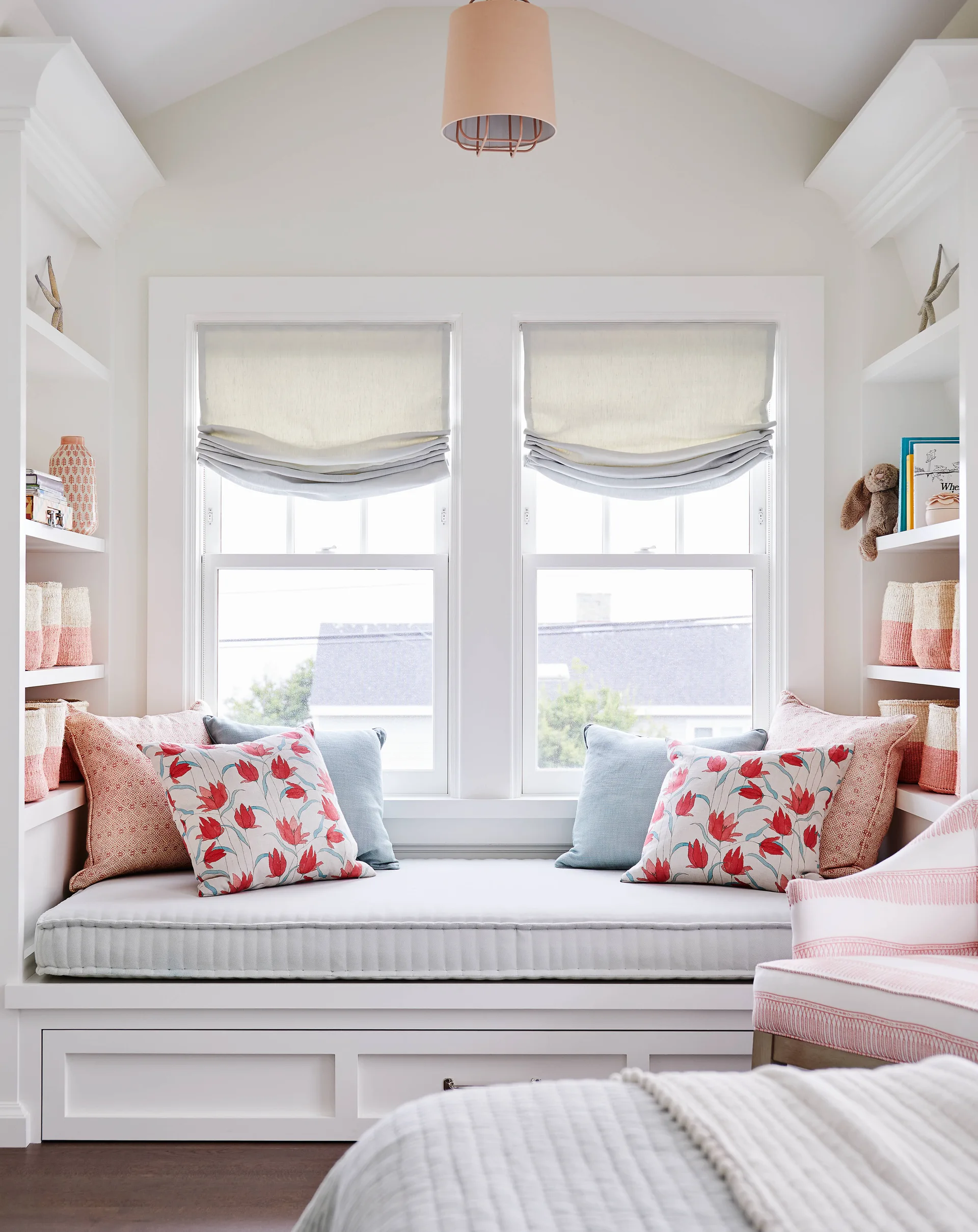 A built-in white window seat with a grey cushion and colorful floral pillows is flanked by symmetrical bookshelves under two windows with roman shades.