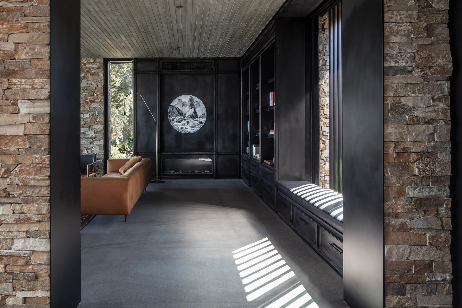 A modern living room features a tan leather sofa, dark matte built-in cabinetry, textured stone walls, and a slatted ceiling.