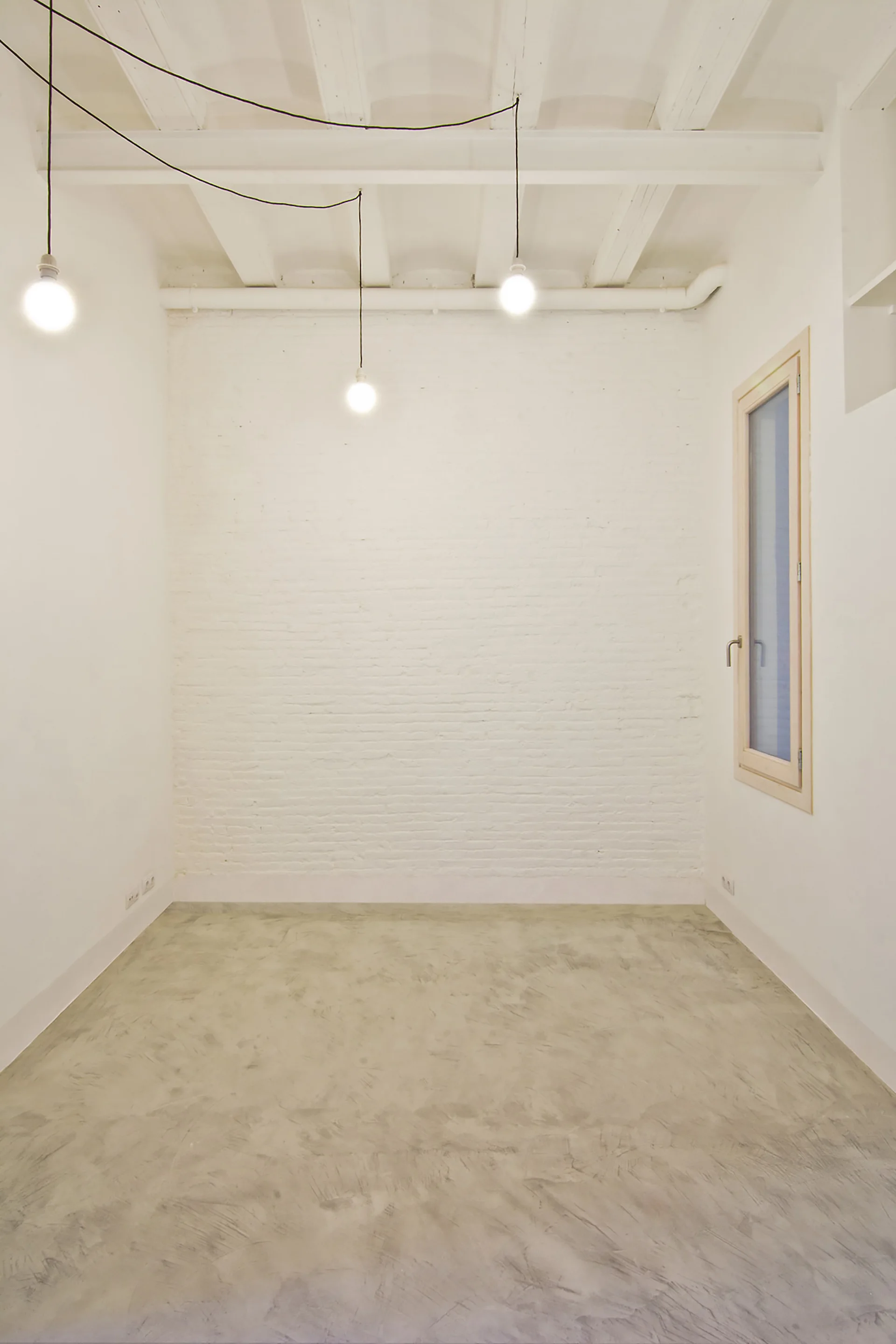 An empty industrial style room with a white painted brick wall, rustic ceiling beams, and bare bulb pendant lights hanging over a gray concrete floor.