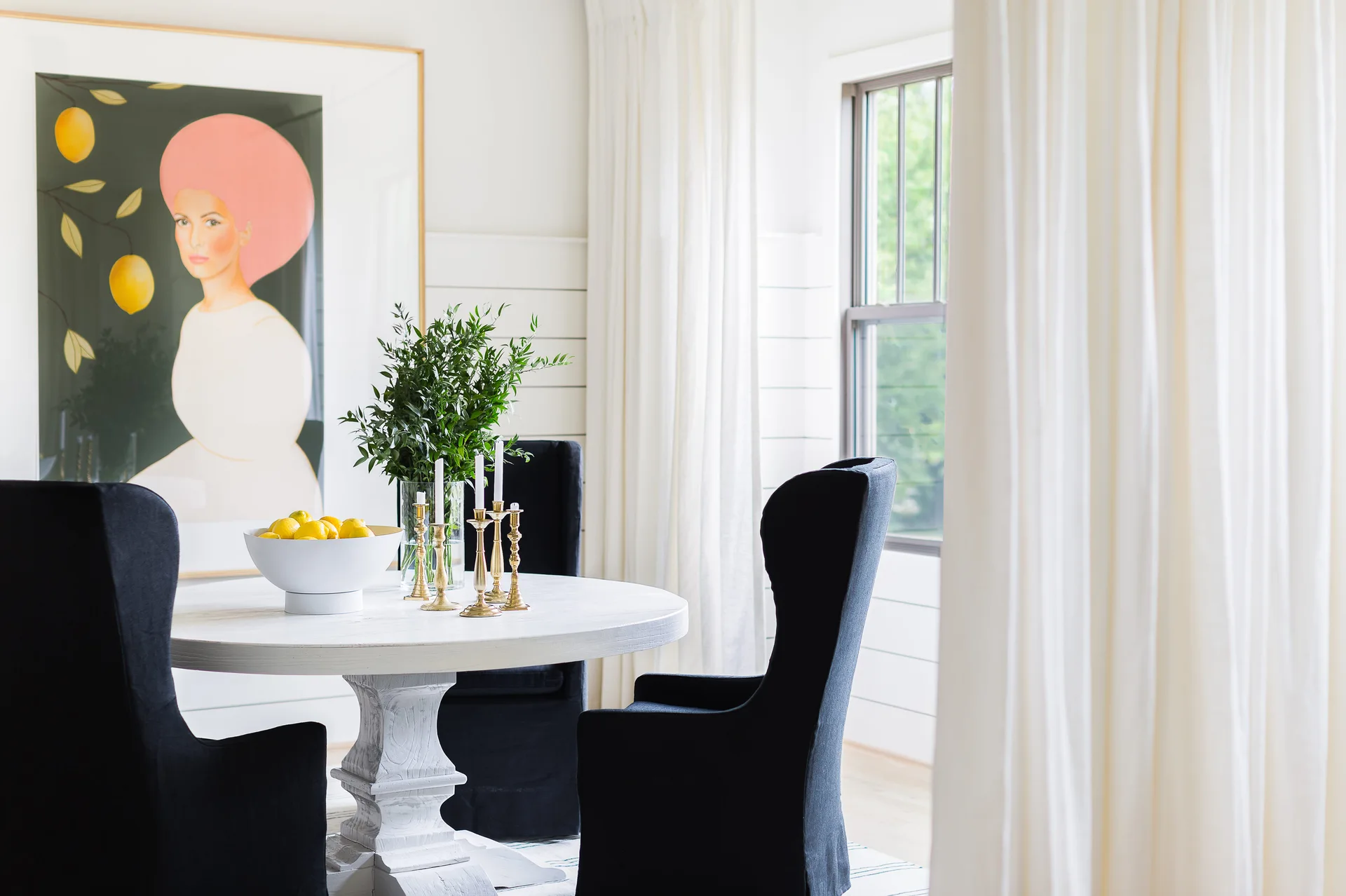 A round white pedestal table with black upholstered chairs sits in a bright room with a shiplap wall and a large framed portrait print.