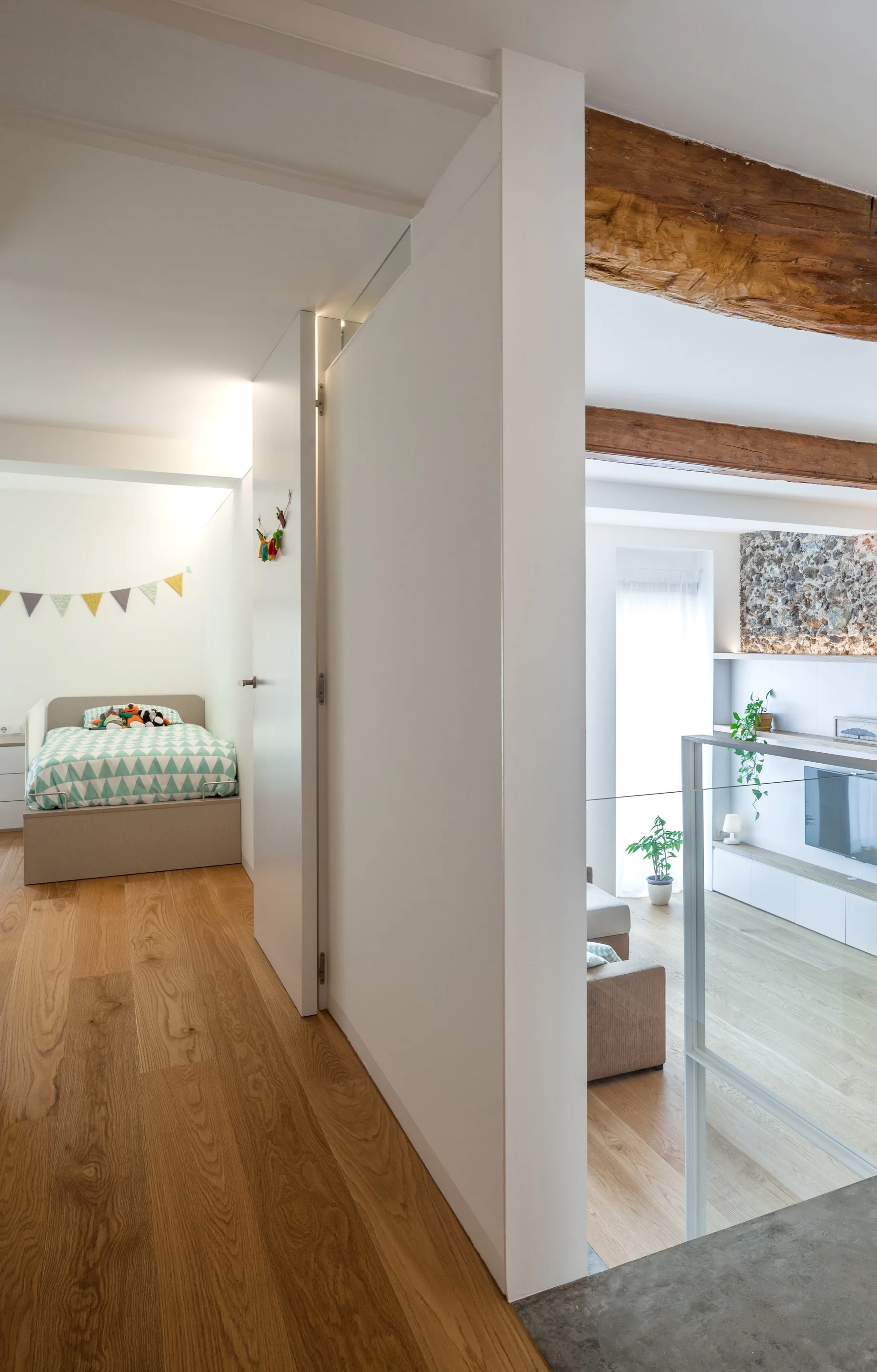 A hallway with wood flooring connects a small bedroom to an open living area featuring rustic ceiling beams, a stone wall, and a glass railing.