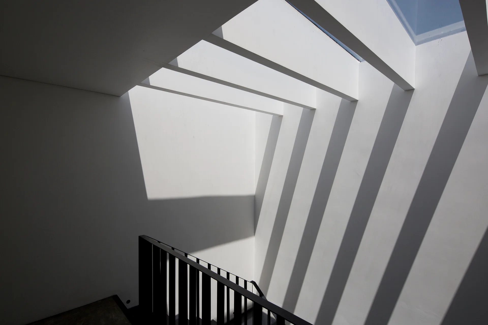 A black metal staircase railing sits below a white ceiling skylight with parallel beams casting bold diagonal shadows on a clean white wall.