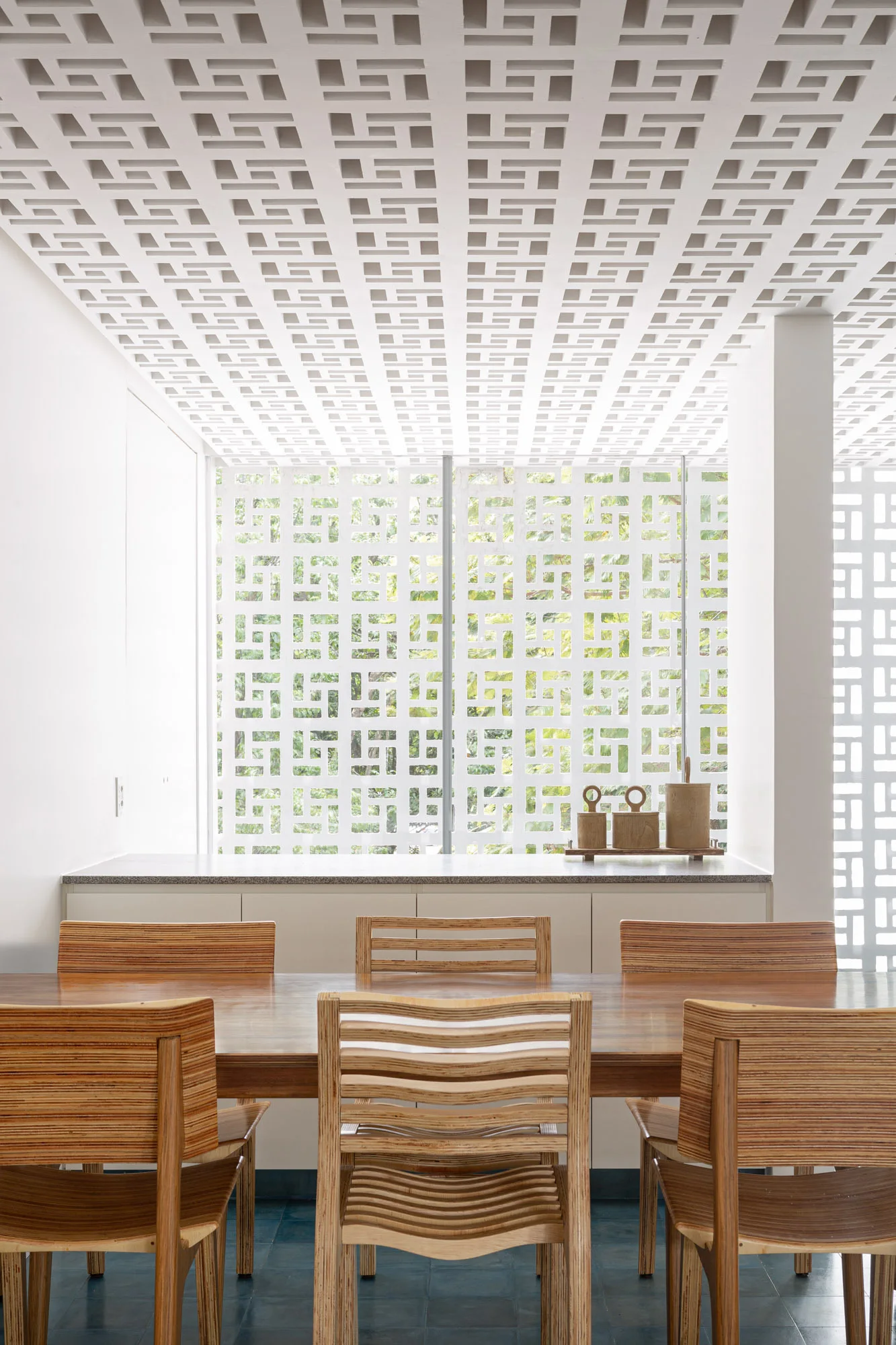 A modern dining room with a long wood table and slatted plywood chairs set against a white geometric breeze block wall and ceiling structure.