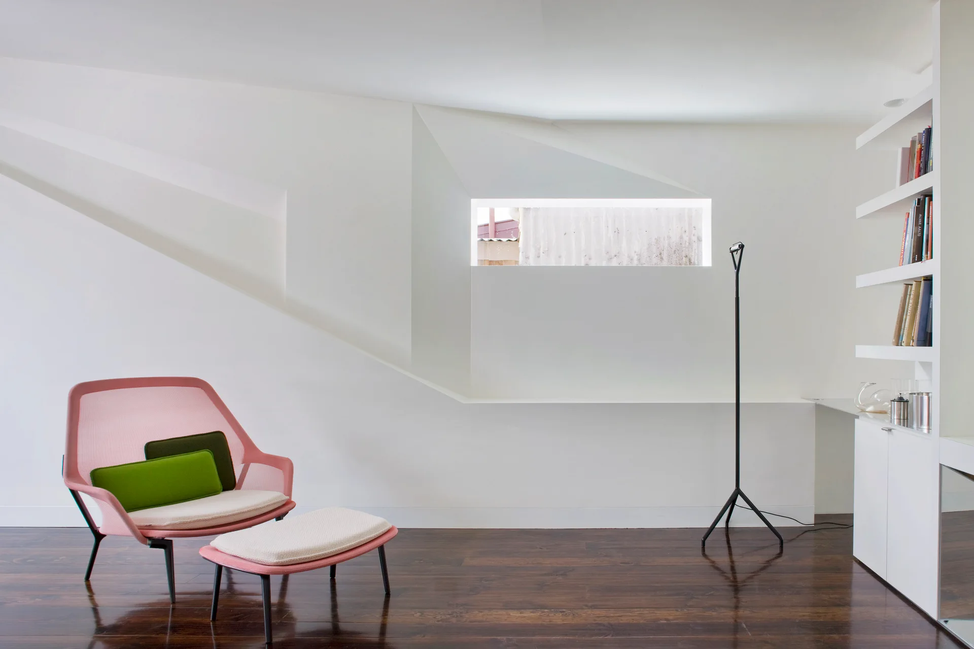 A pink mesh lounge chair and matching ottoman sit on dark wood floors in a minimalist white room with an angled wall cutout.