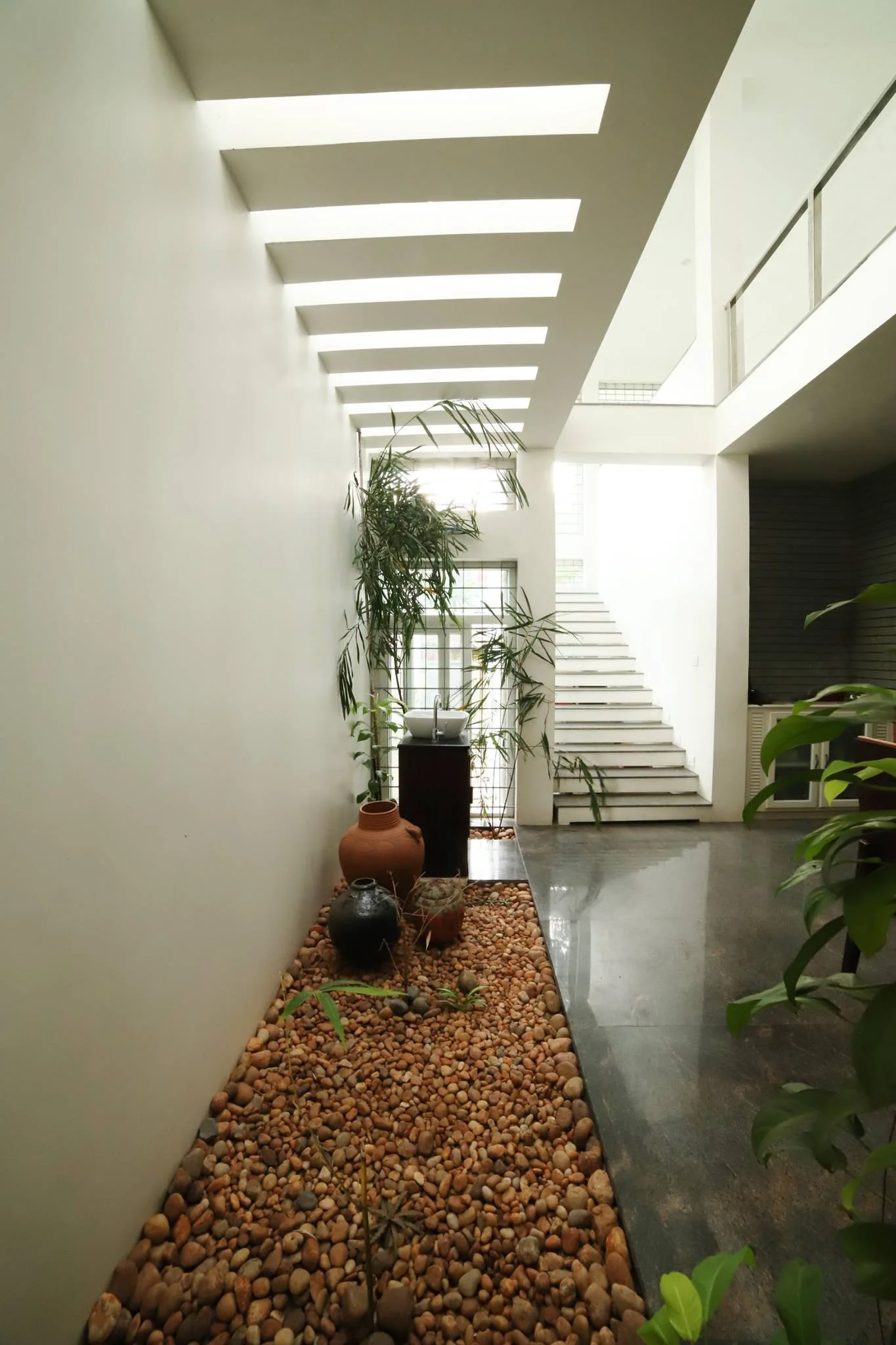 A modern entryway hallway with a pebble-filled indoor garden, earthenware pots, a slatted skylight overhead, and a white staircase.