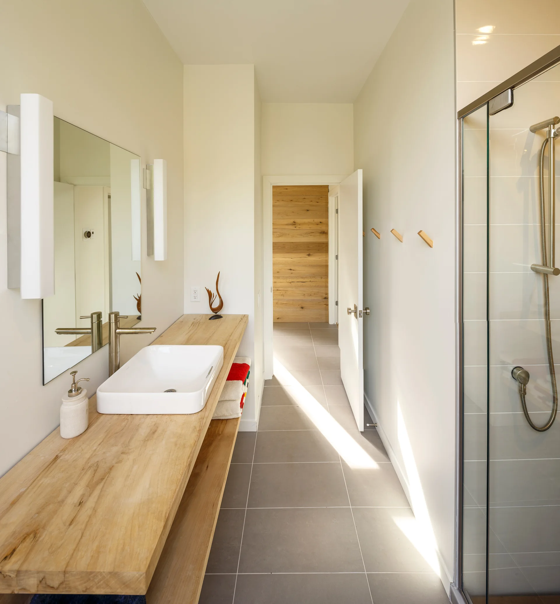 A modern bathroom with a long natural wood slab vanity holding a white vessel sink, set against light grey floor tiles and a glass-enclosed shower.