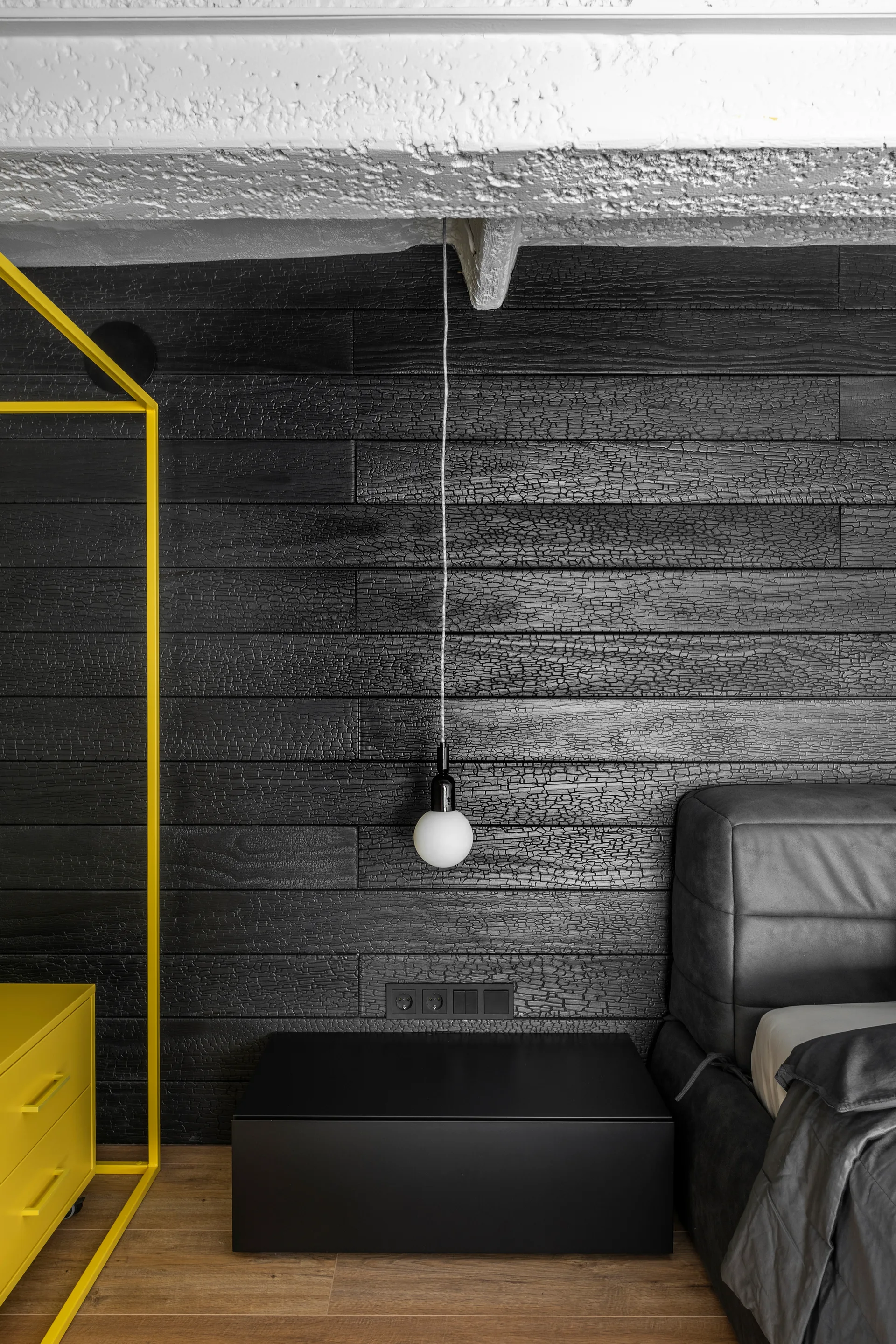 A modern bedroom features a textured black charred wood wall, a low black nightstand, a hanging globe pendant, and a bright yellow frame.