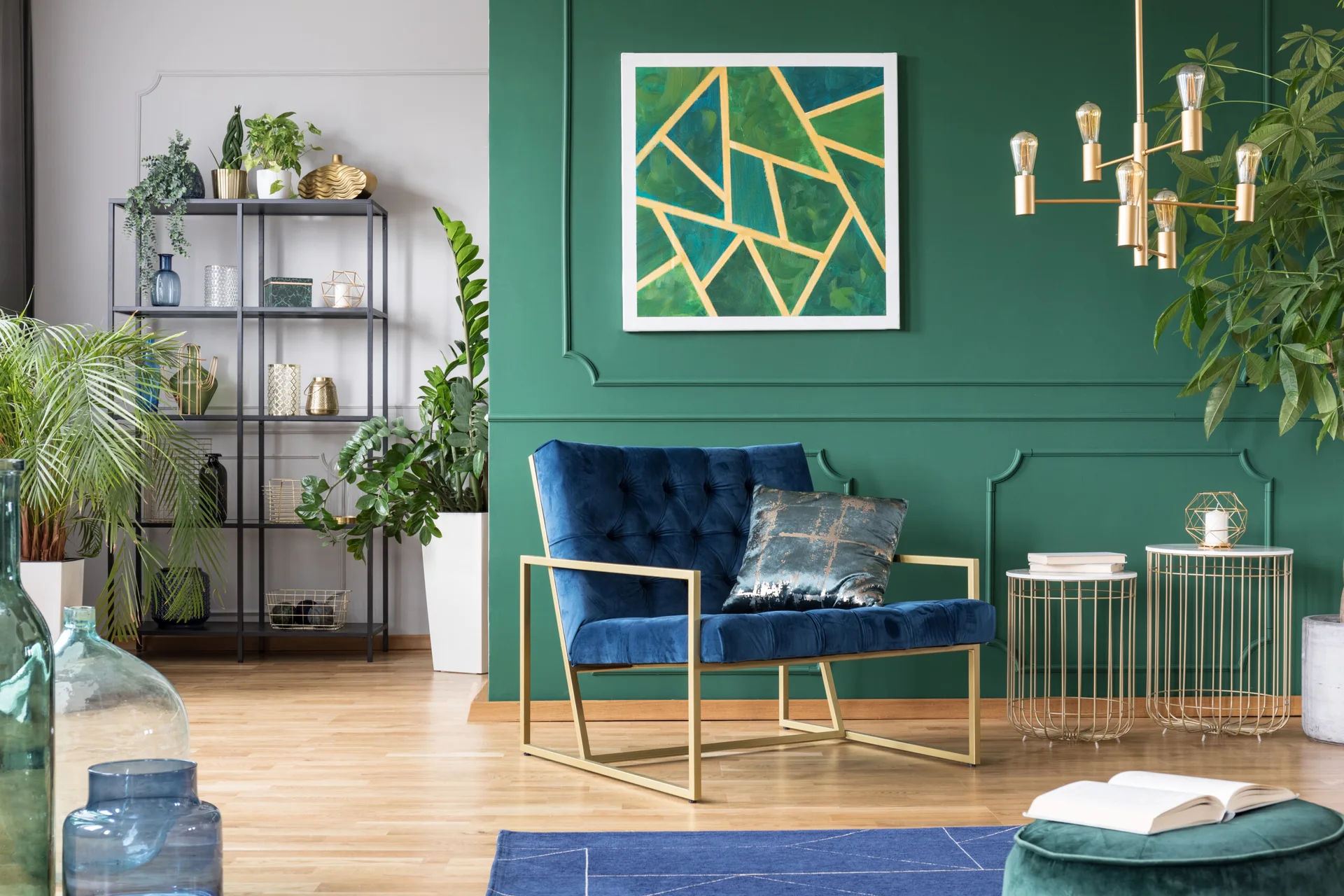 A royal blue velvet armchair with a gold frame sits in front of a forest green wall featuring decorative moulding and geometric art.