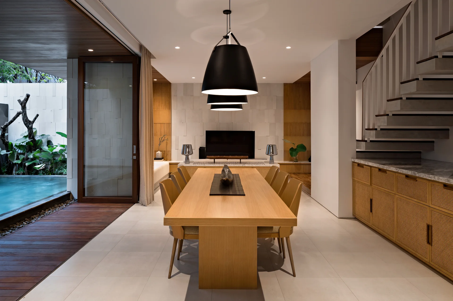 A minimalist dining room features a long wooden table, black dome pendant lights, and cane cabinets next to a floating staircase.