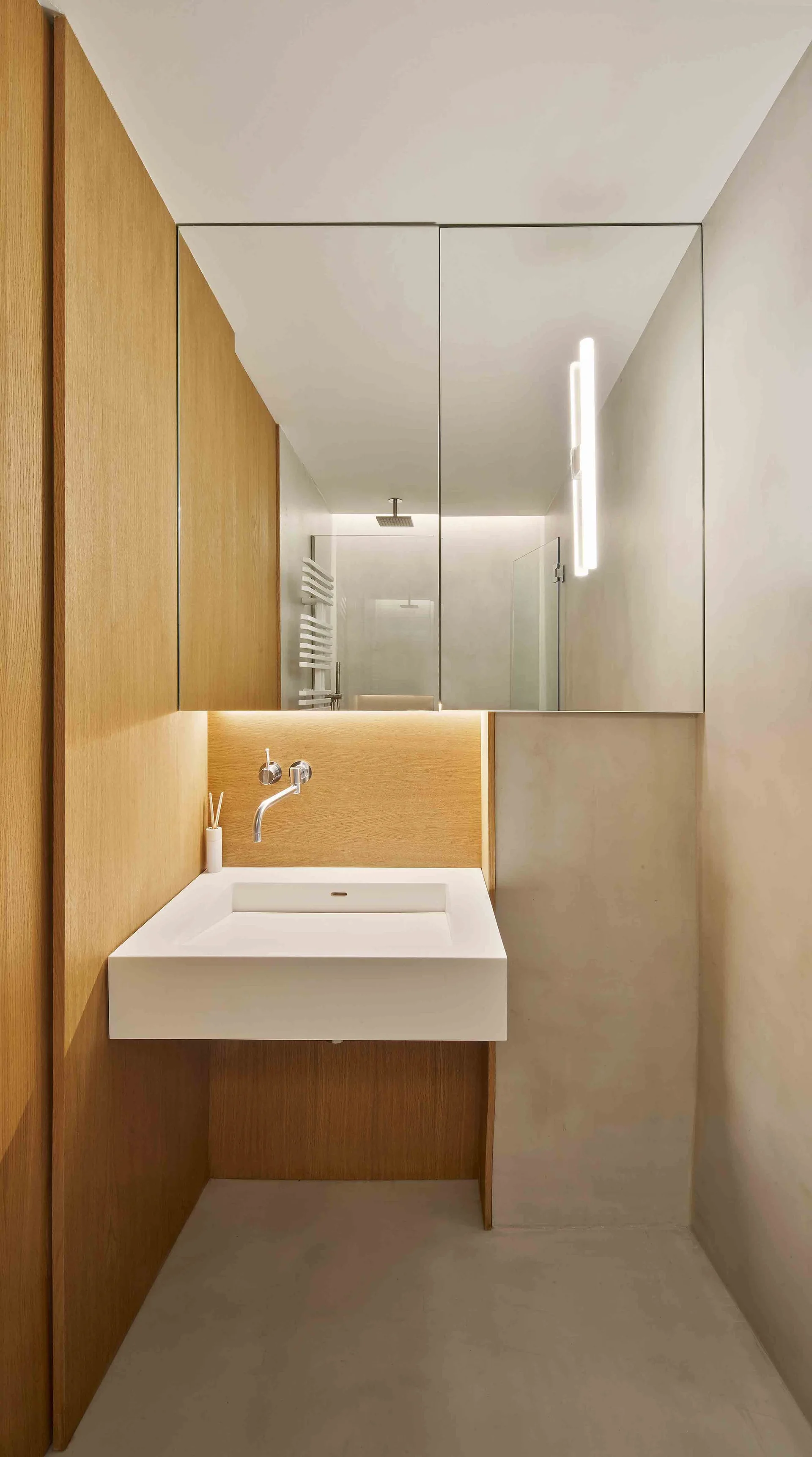 A minimalist bathroom features a floating white vanity sink against warm wood paneling, a concrete wall, and a large mirror cabinet.