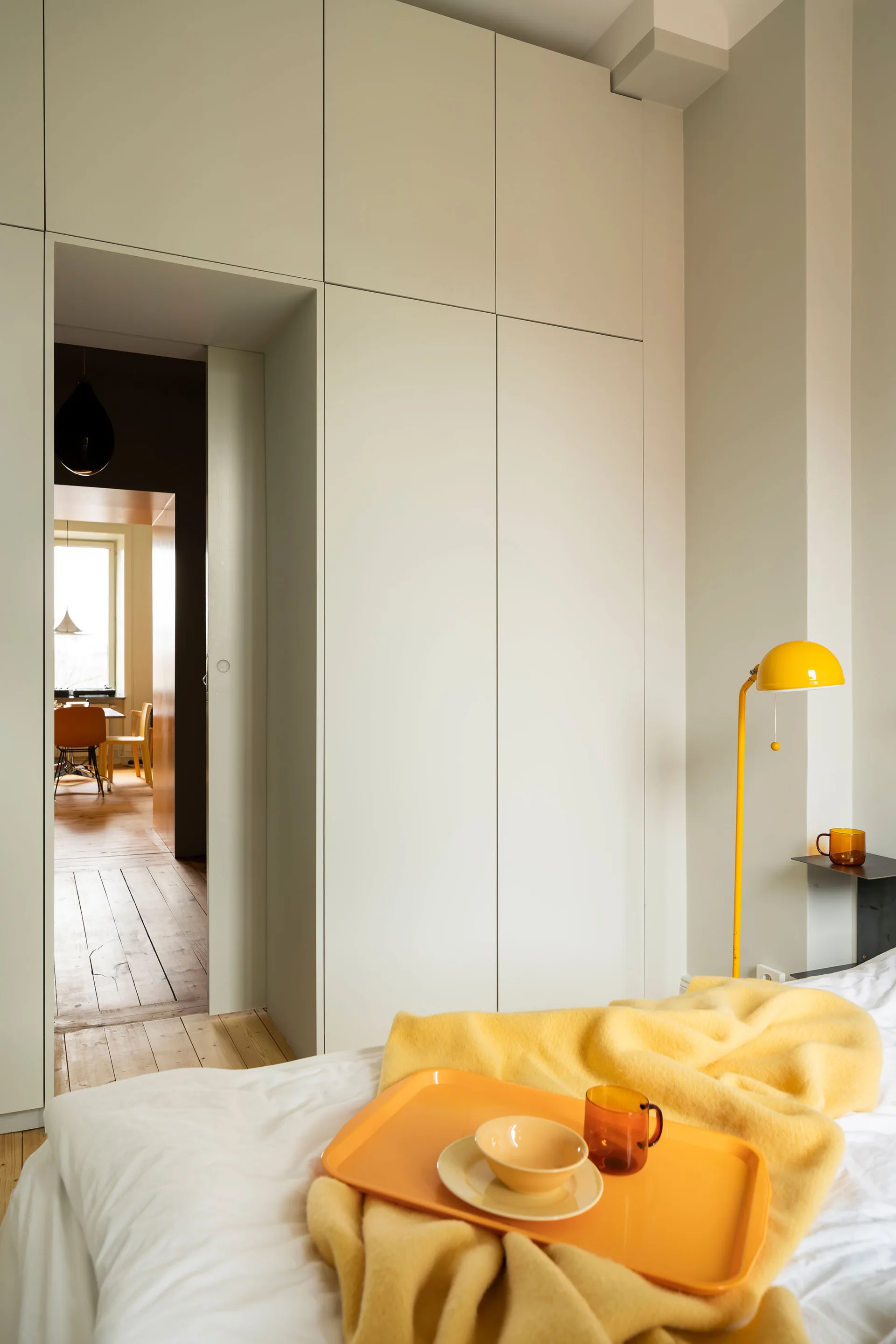 A minimalist bedroom features seamless off-white built-in cabinetry, a bed with a yellow throw and tray, and a yellow floor lamp next to a doorway.