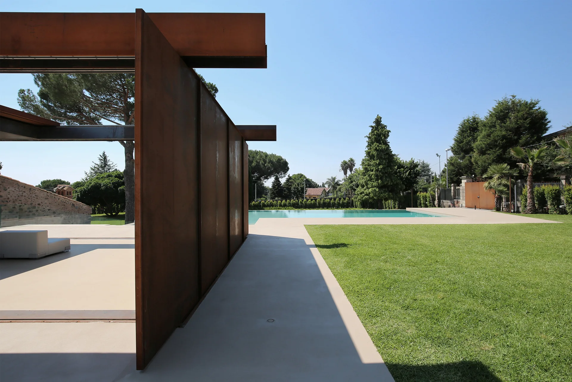 A modern outdoor swimming pool is surrounded by a manicured green lawn, concrete patio, and a prominent weathered rust-colored metal wall structure.