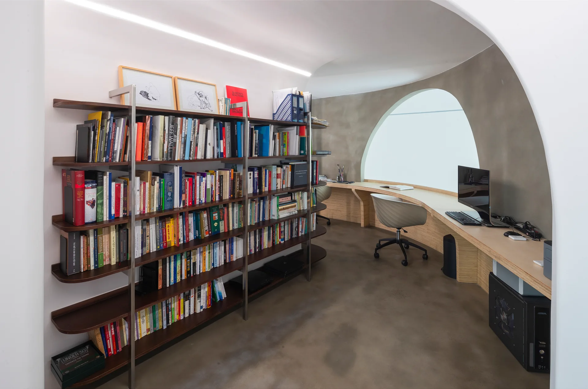 A modern home office featuring a curved wooden desk built along a grey wall, a large bookshelf filled with books, and an arched window.