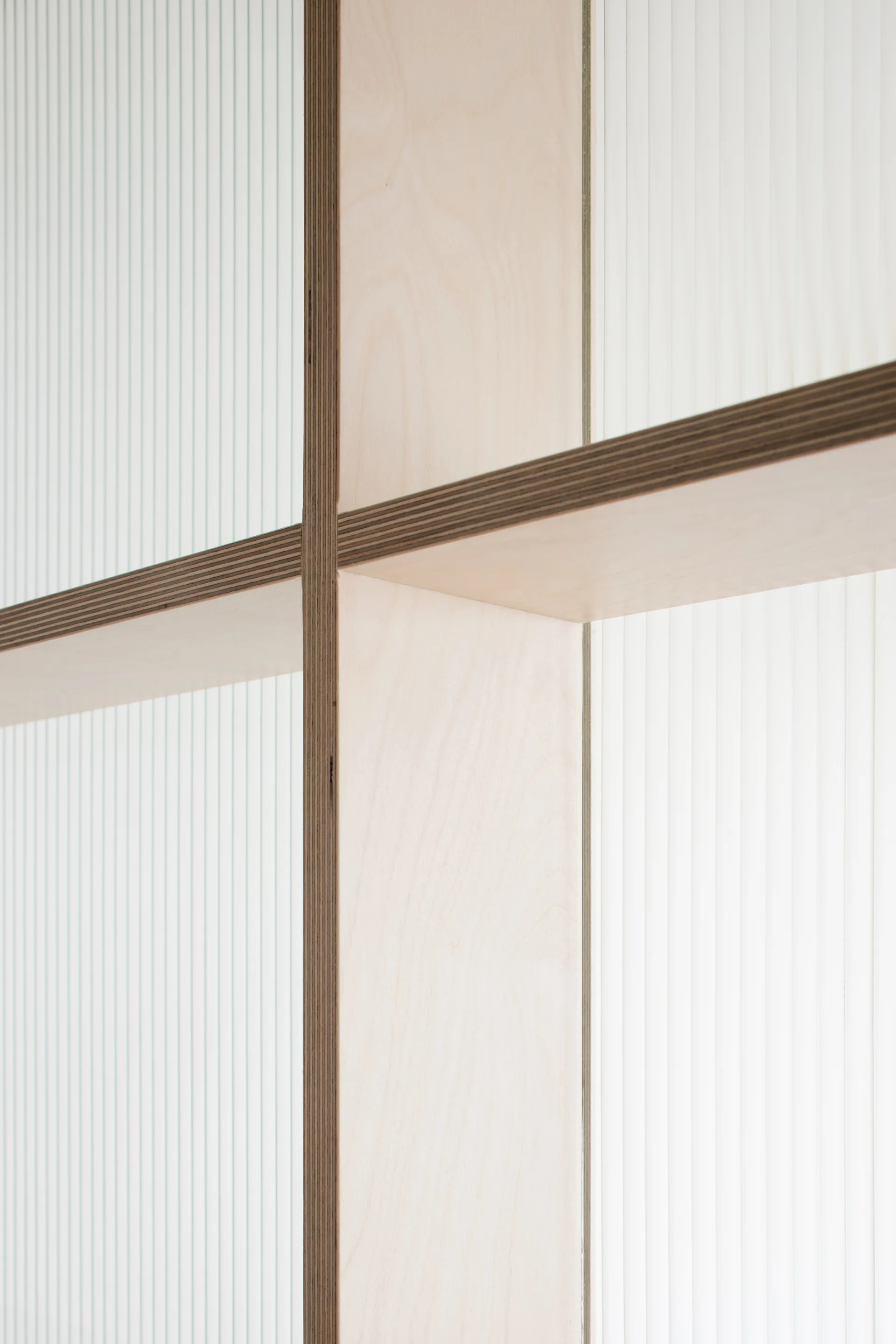 A close-up view of a light-colored plywood frame intersection set against a translucent vertically ribbed fluted glass panel.