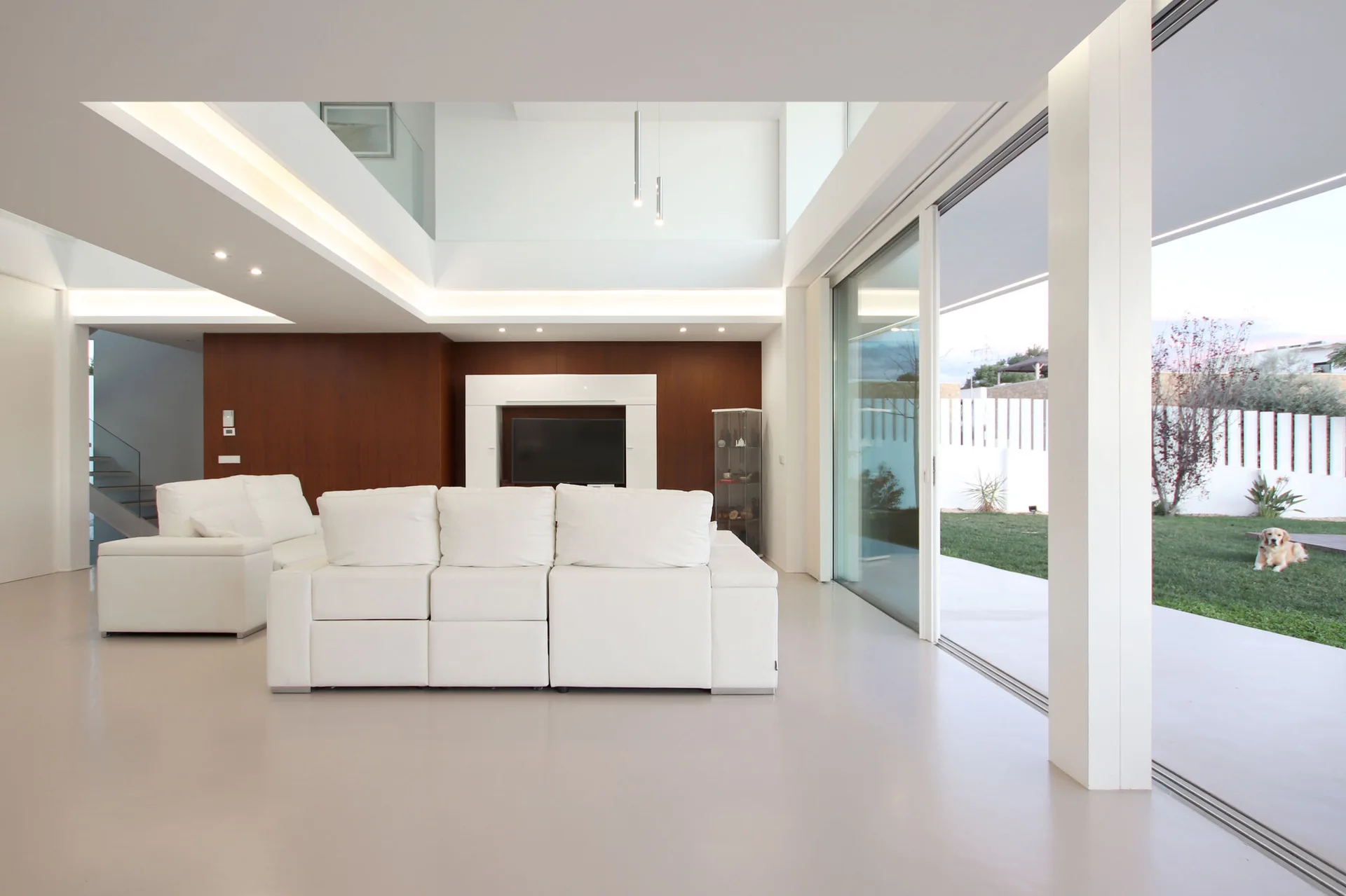 A minimalist white living room features a large white sectional sofa, a dark wood-paneled accent wall, and sliding glass doors opening to a yard.