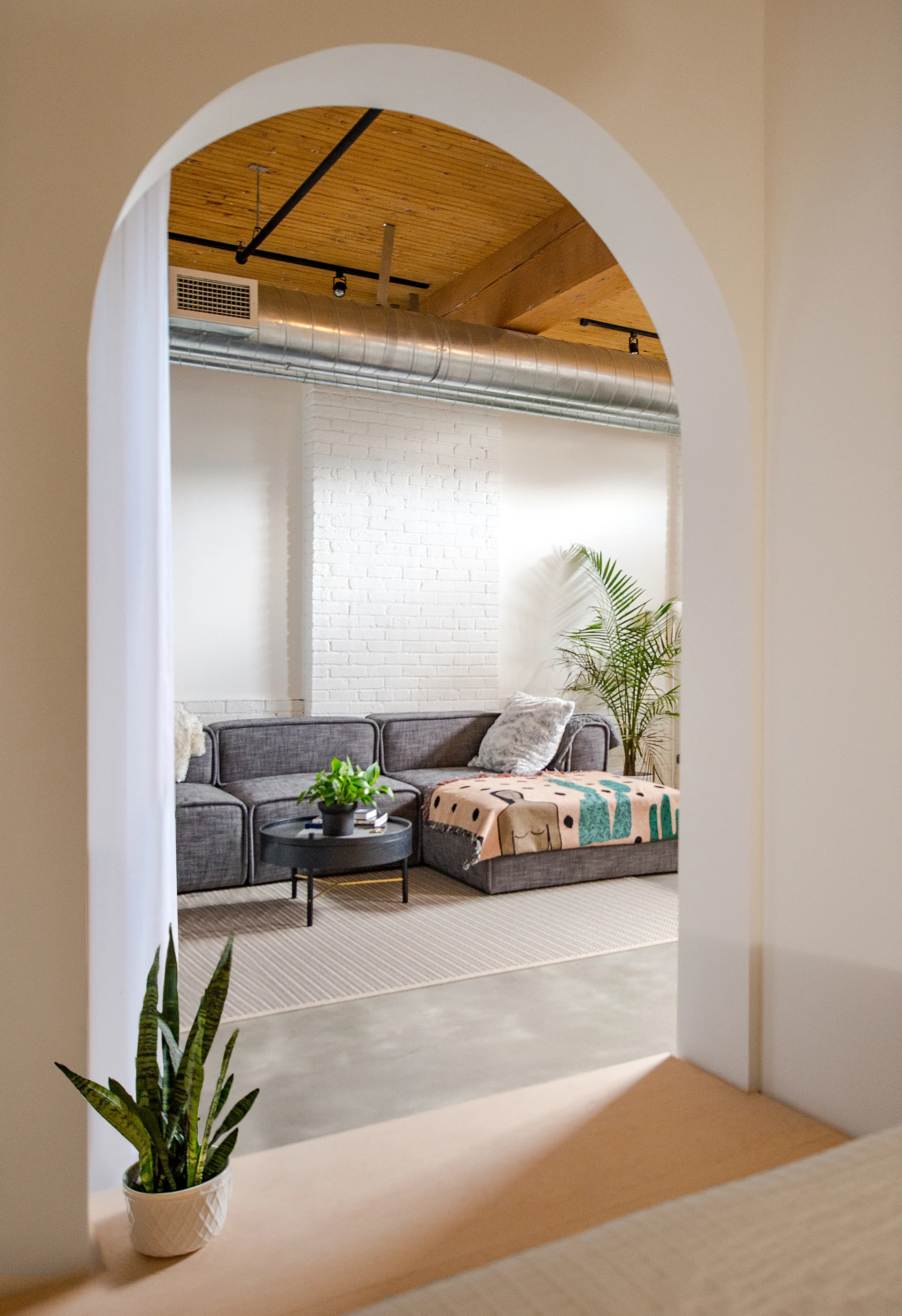 A view through a white arched doorway showing a grey sectional sofa, round coffee table, white brick wall, and exposed wooden ceiling beams.