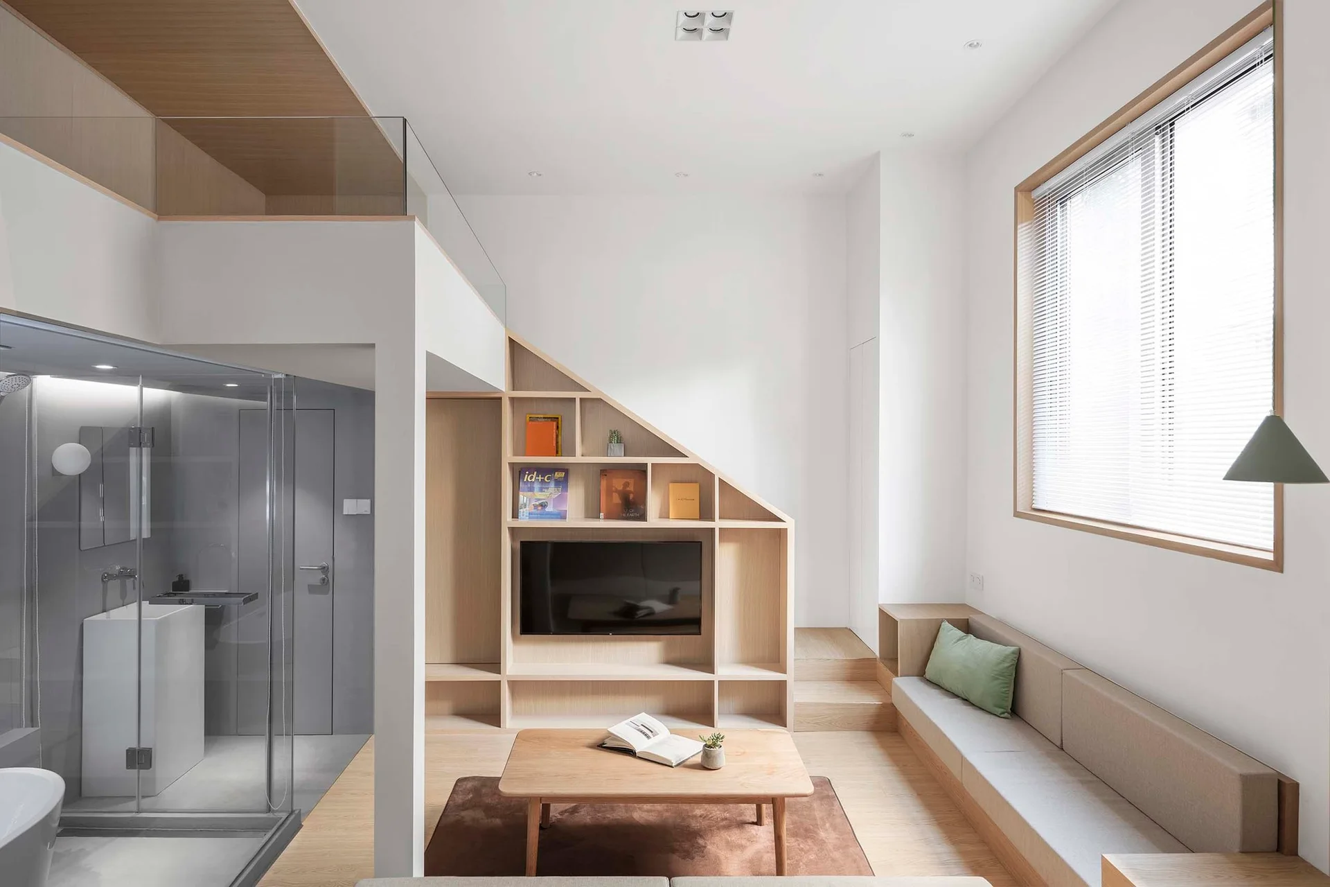 A minimalist loft living room features a custom sloped light wood shelving unit with a television, a built-in upholstered bench, and a glass partition.