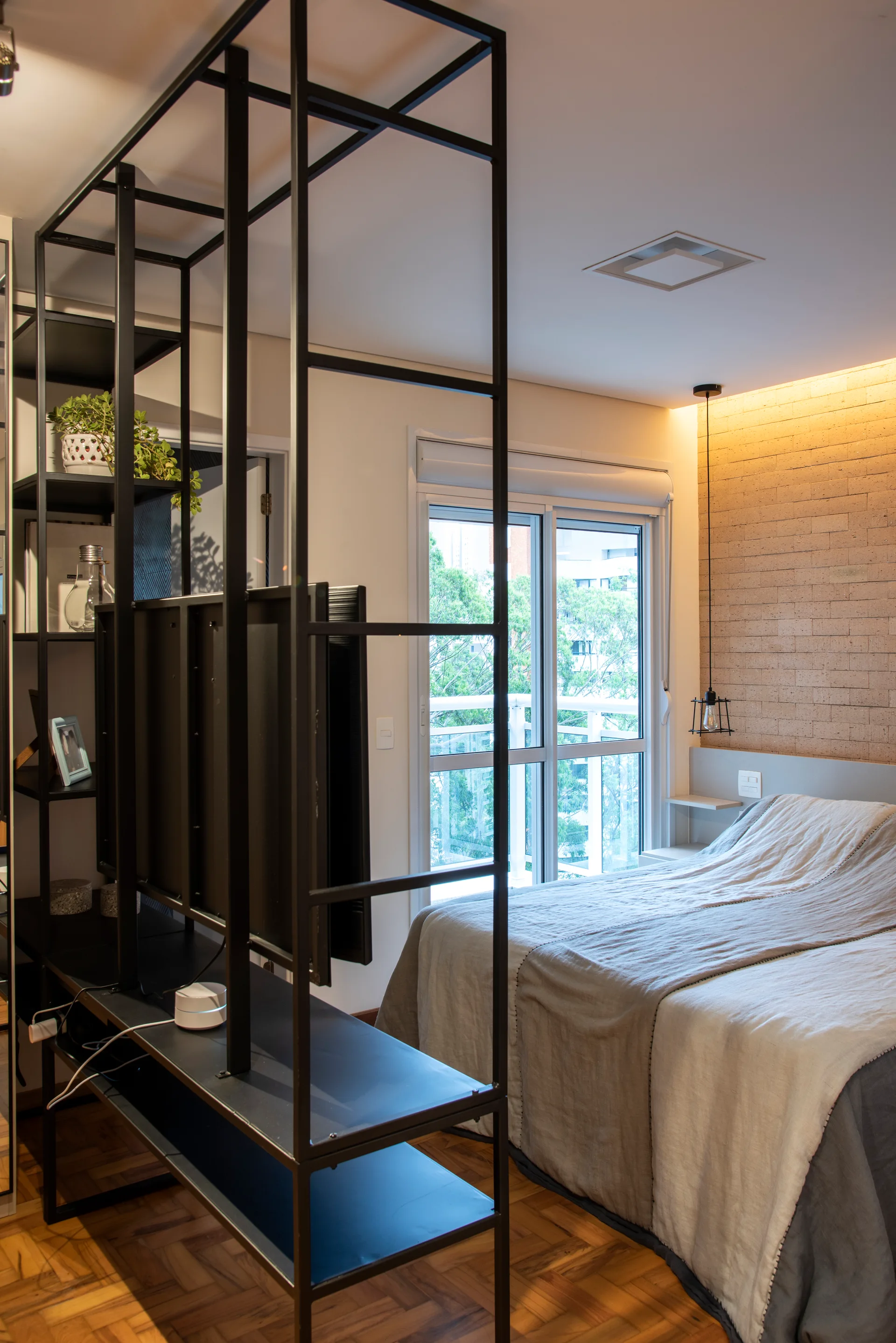 A black metal frame room divider with a mounted TV stands near a bed with grey linens in a bedroom with a brick feature wall and sliding glass door.