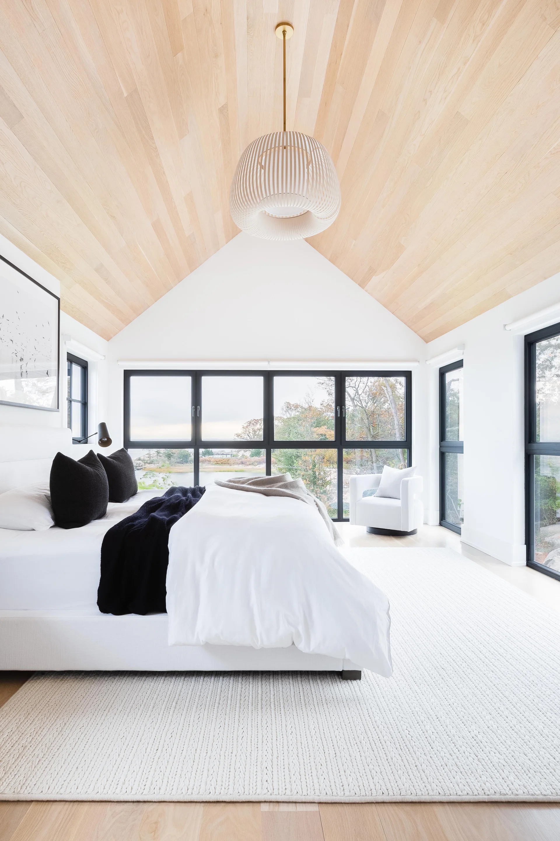 A modern bedroom features a vaulted light wood ceiling, a white bed with black pillows, and large black-framed windows looking out to trees.
