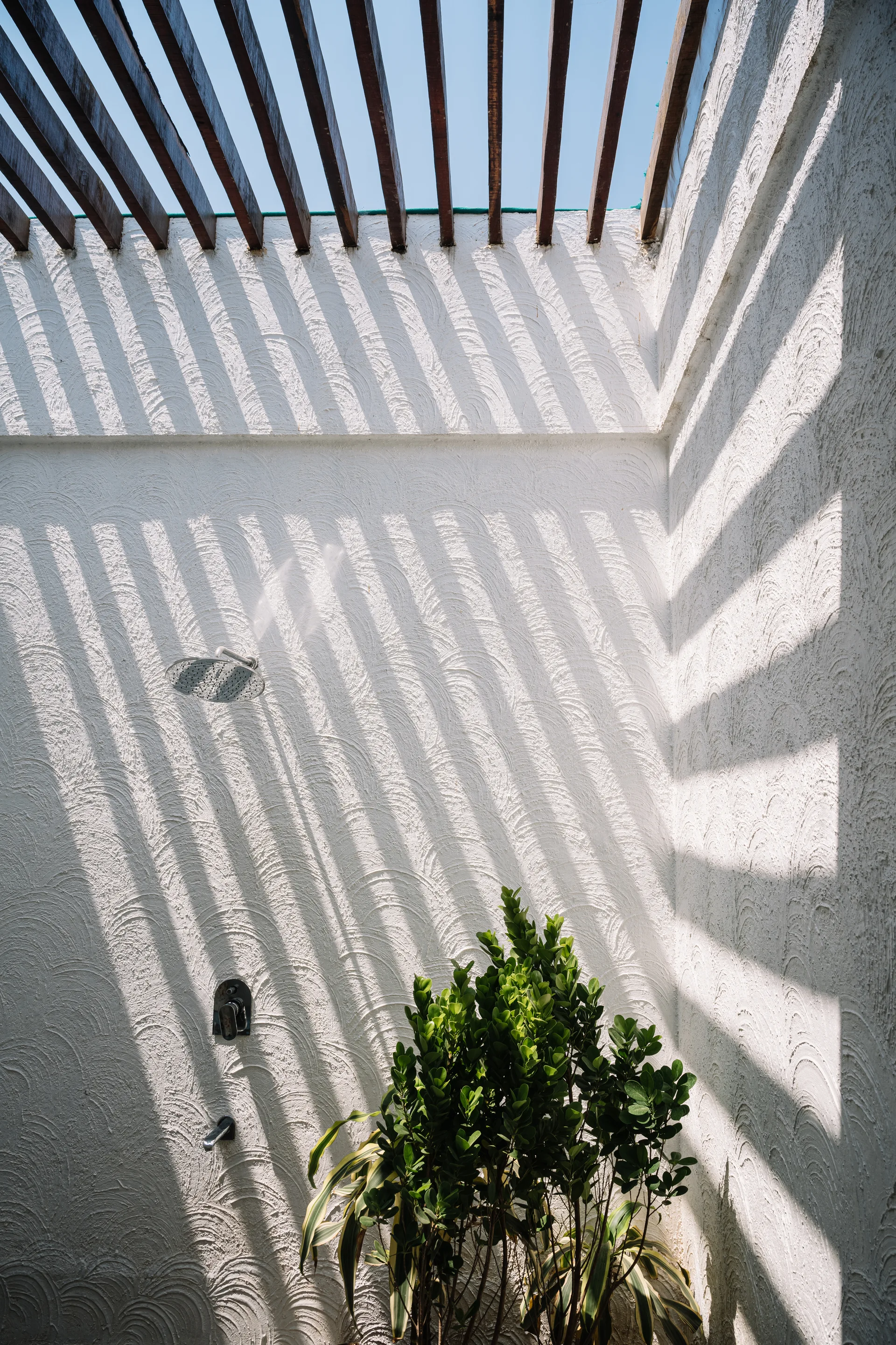 An outdoor shower with a metal shower head and faucet features a textured white plaster wall under a slatted wooden pergola that casts linear shadows.