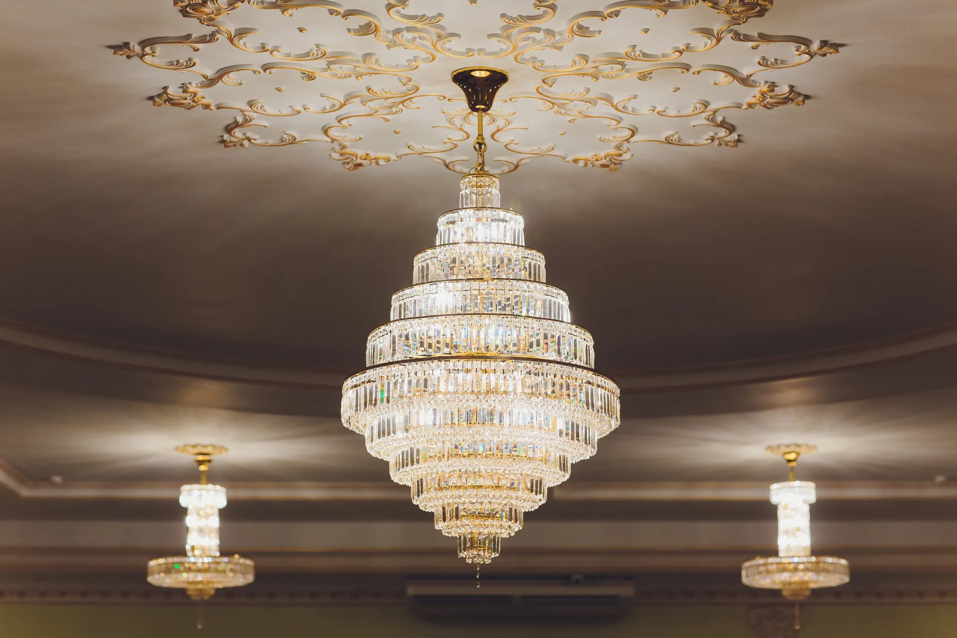 A tiered crystal chandelier hangs from a white ceiling adorned with ornate gold plasterwork scrolls, with secondary fixtures visible in the background.
