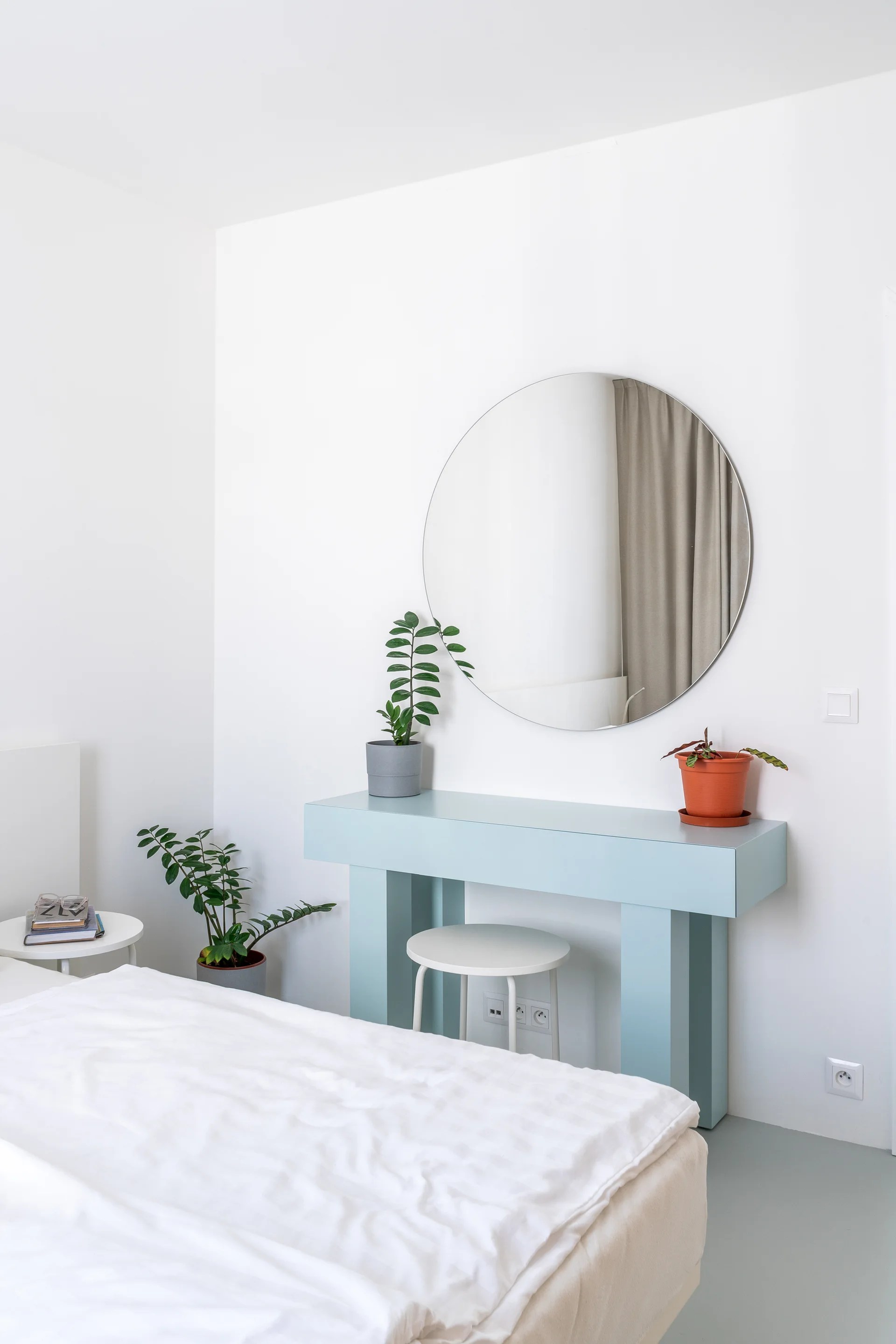 A minimalist white bedroom featuring a pale blue vanity table with a round mirror, small potted plants, and white bedding.