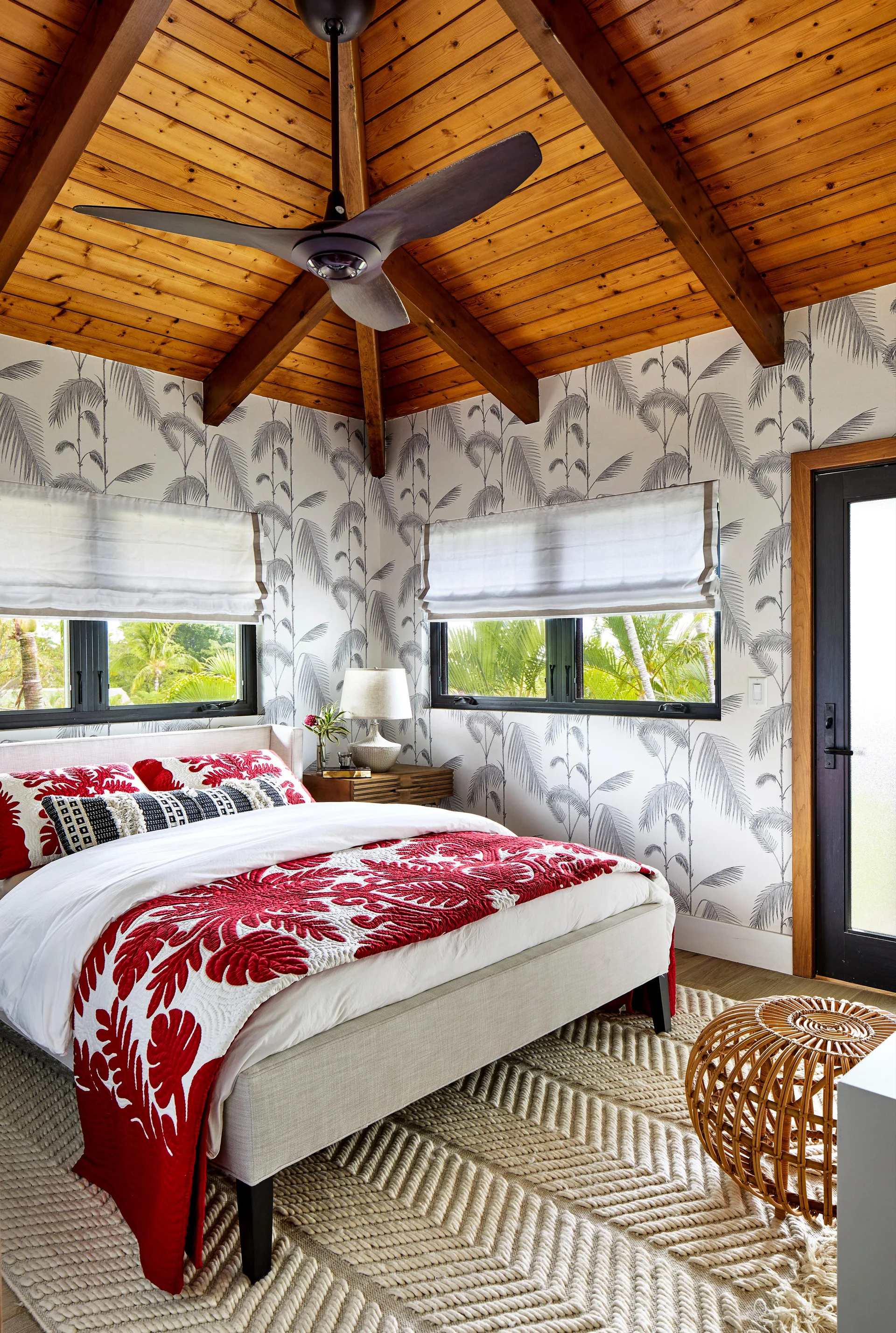 A tropical bedroom with a vaulted wood ceiling, black-and-white palm wallpaper, a bed with a red quilt, and a textured chevron rug.