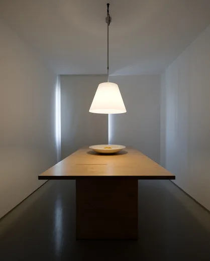 A minimalist dining room features a centered rectangular wooden table beneath a glowing tapered pendant light against smooth white walls.