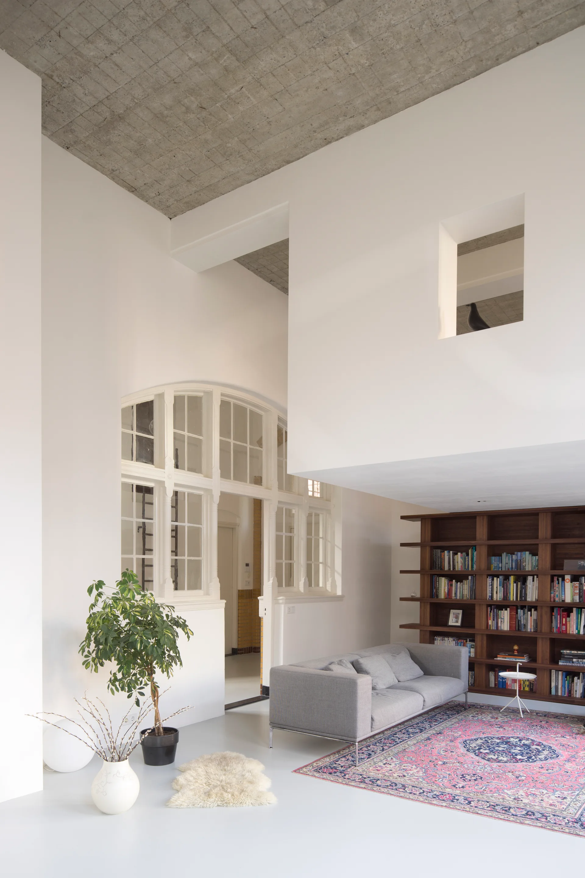 A spacious loft living room features a grey sofa, a dark wood bookcase, and a pink patterned rug beneath an exposed concrete ceiling and arched partition.