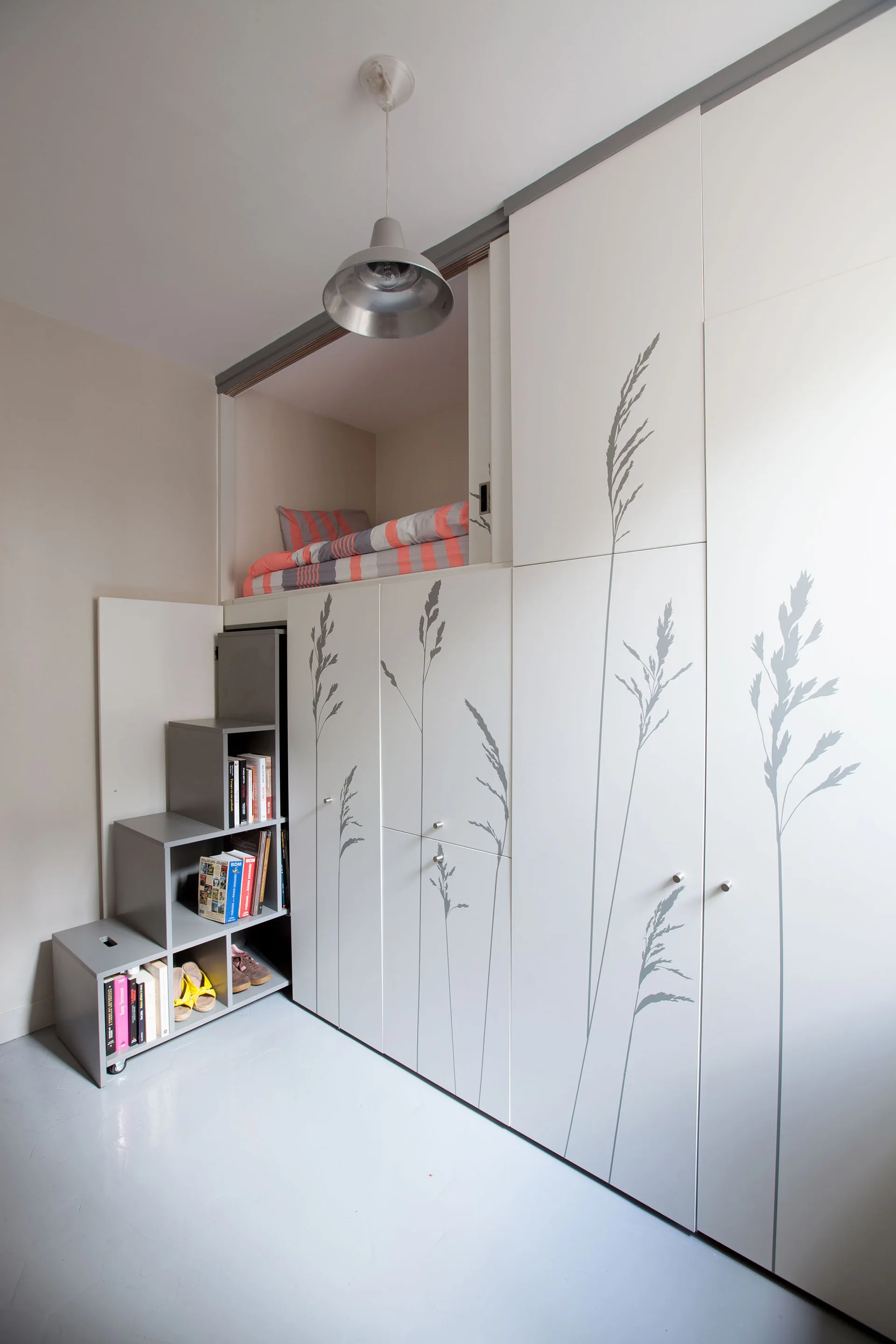 A compact bedroom featuring a loft bed over white built-in wardrobes with grey grass graphics and a mobile grey step shelf holding books.