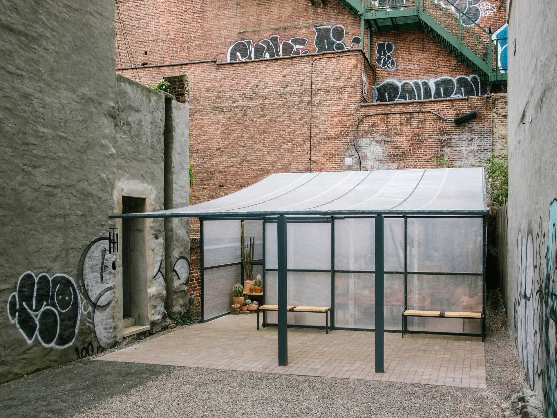 An outdoor courtyard features a translucent plastic-roofed shelter with metal frames, wooden benches, and potted cacti against graffiti-covered brick walls.