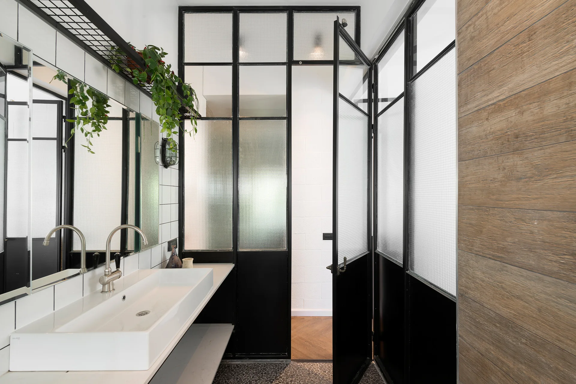 A modern bathroom features a white trough sink, a black metal grid glass partition, and a wood-paneled wall.