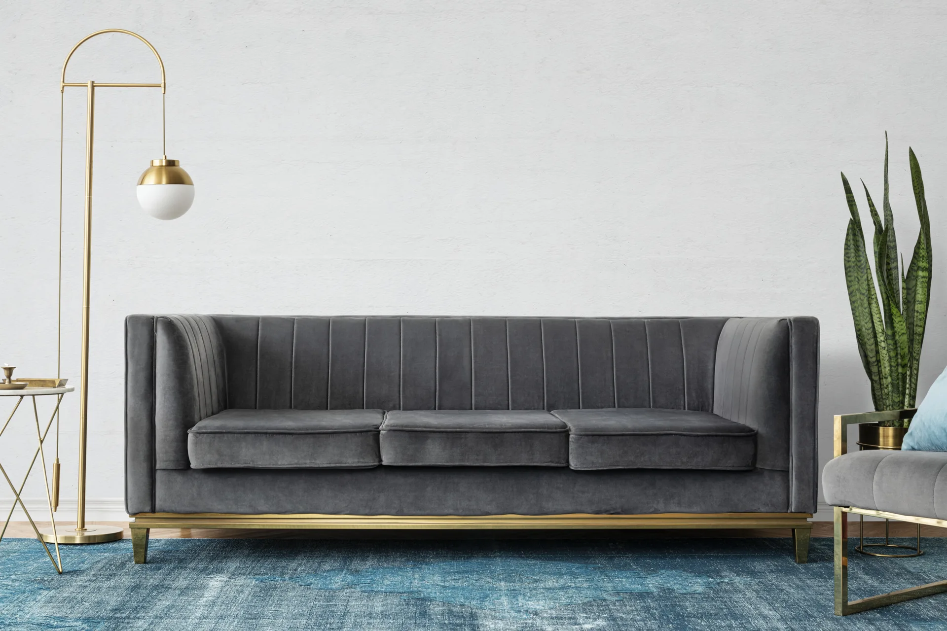 A grey channel-tufted velvet sofa rests on a blue textured area rug next to a gold floor lamp and a tall snake plant.