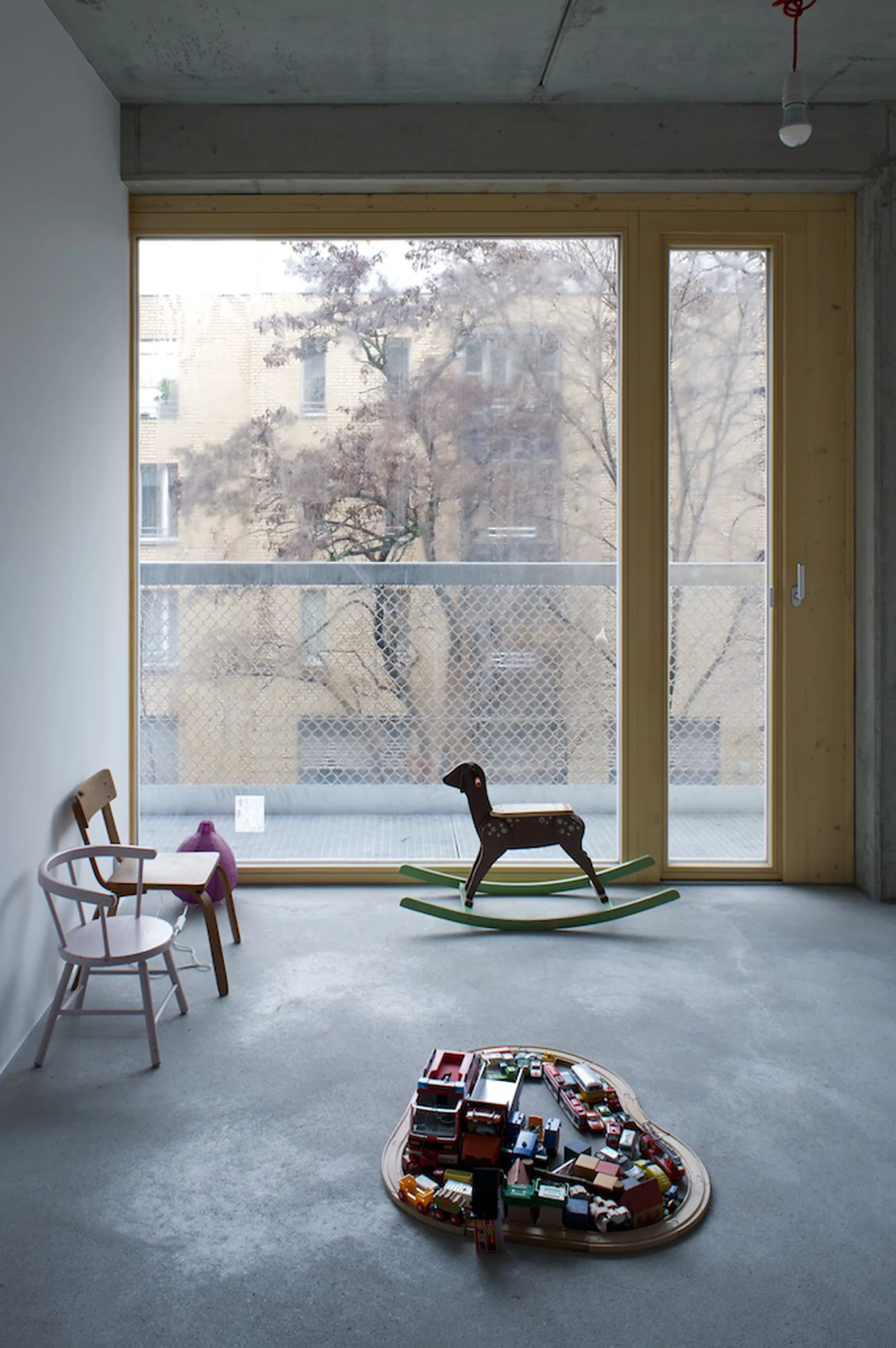 A minimalist children's playroom features a concrete floor and ceiling, a large wood-framed window, a wooden rocking horse, and small chairs.