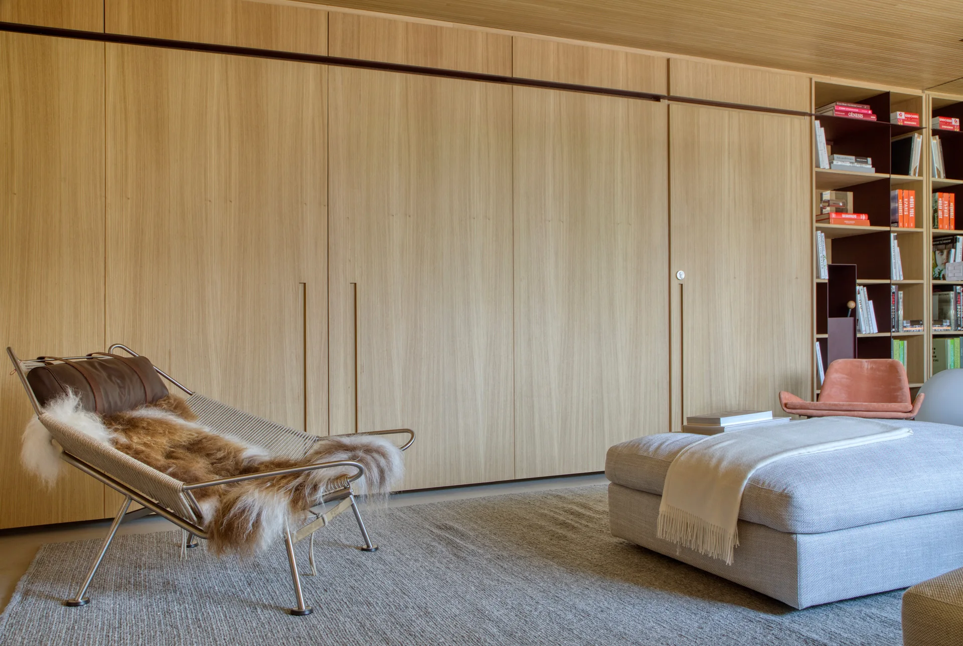A modern living room features floor-to-ceiling wood wall paneling, built-in bookshelves, a metal-framed lounge chair with a fur throw, and an ottoman.