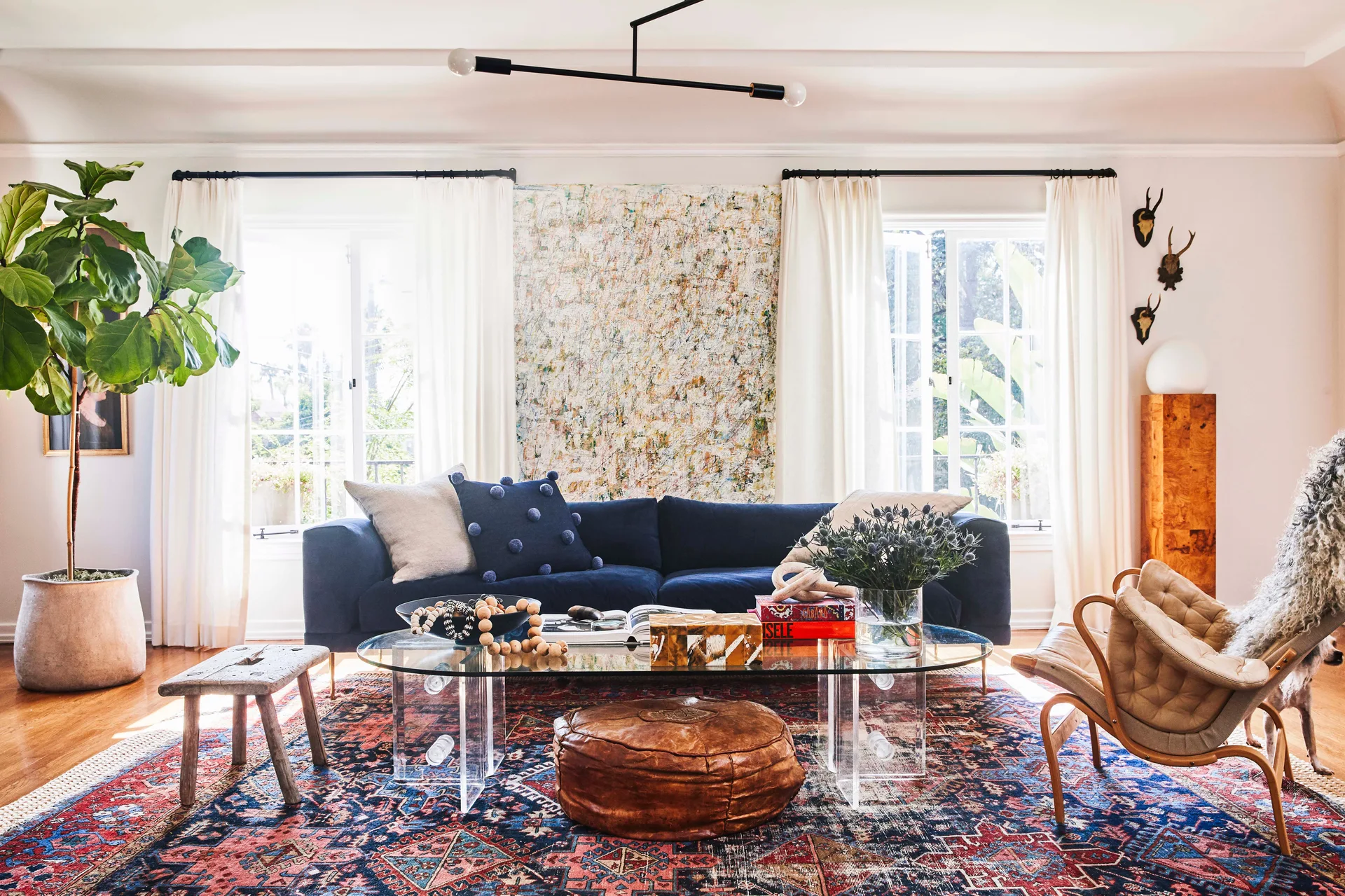 A navy velvet sofa sits on a patterned oriental rug in a living room with an oval glass coffee table, abstract art, and two curtained windows.
