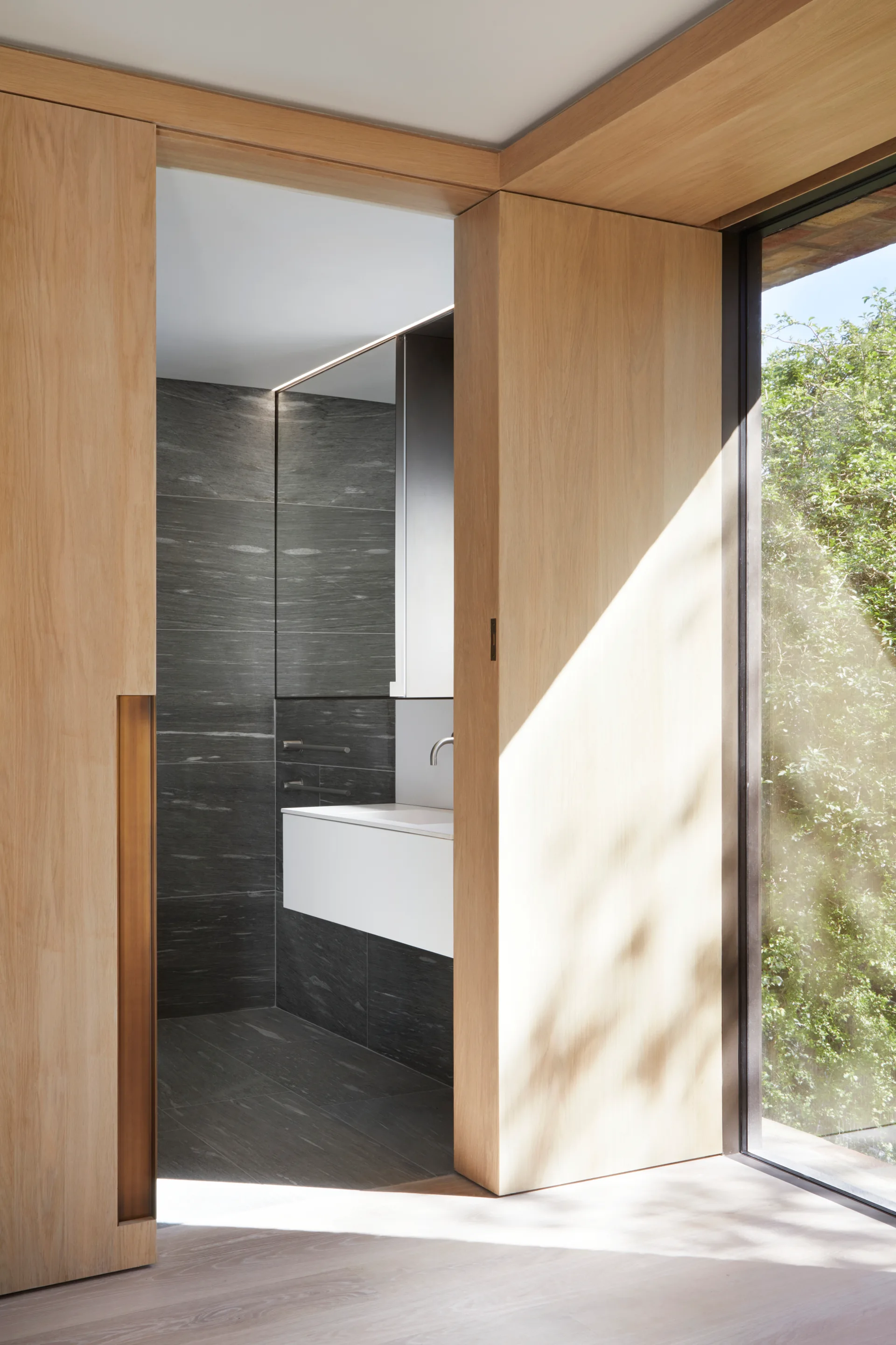 A light wood pocket doorway opens to a modern bathroom featuring dark gray stone wall tiles and a floating white vanity.