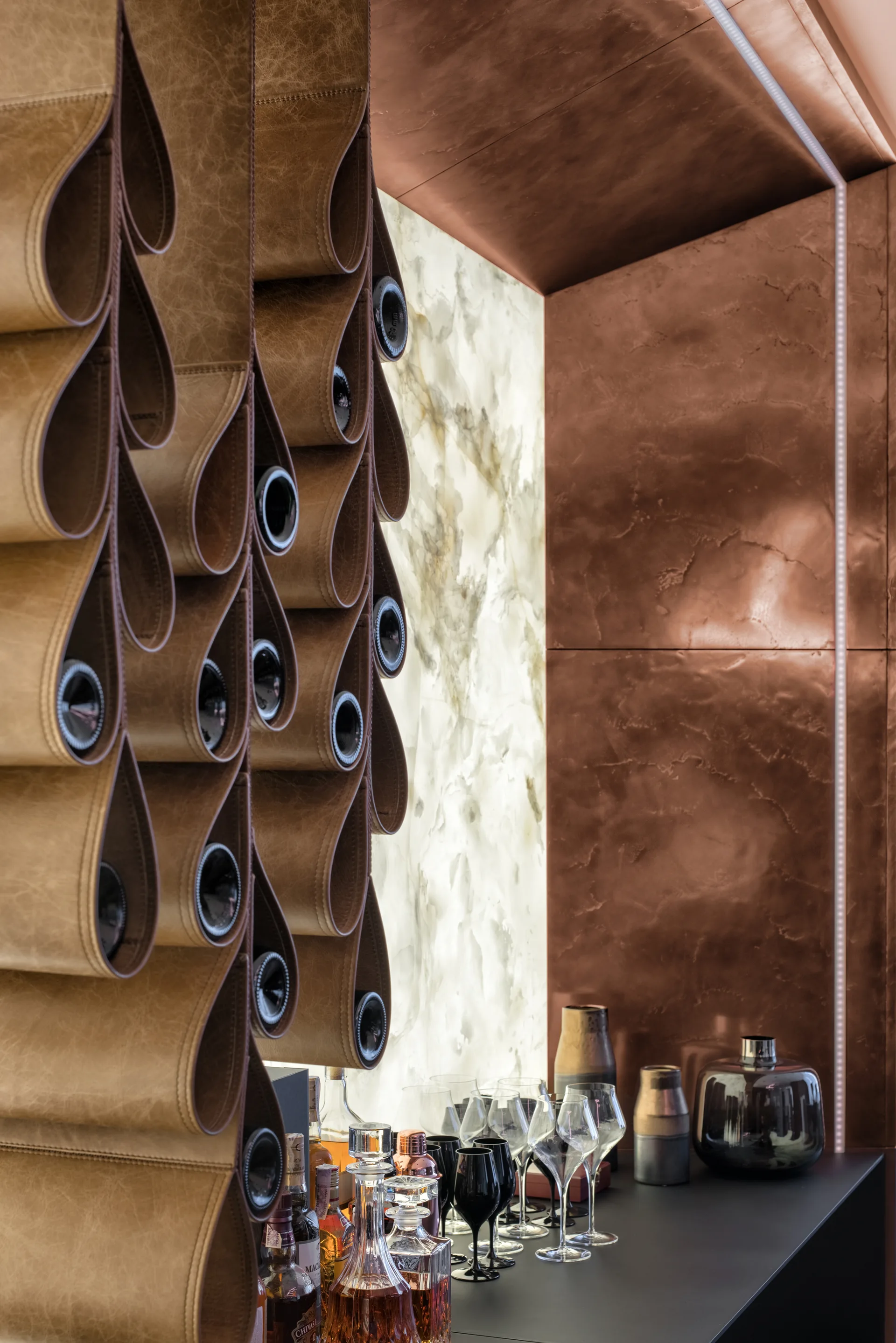 A custom leather loop wine rack hangs next to a glowing backlit marble slab wall above a black home bar counter with glasses.