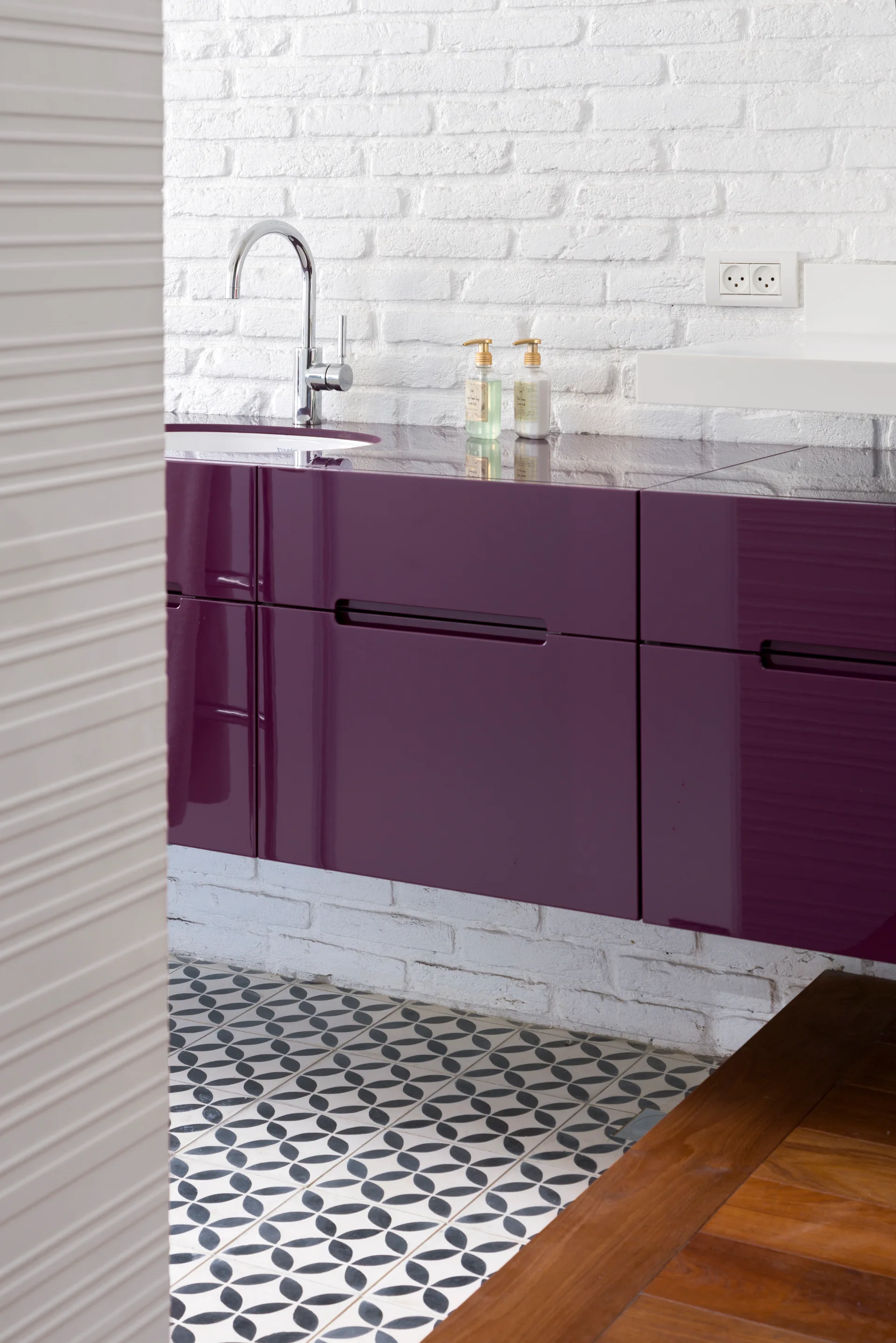 A floating high-gloss purple bathroom vanity is mounted against a white painted brick wall with geometric patterned floor tiles below.