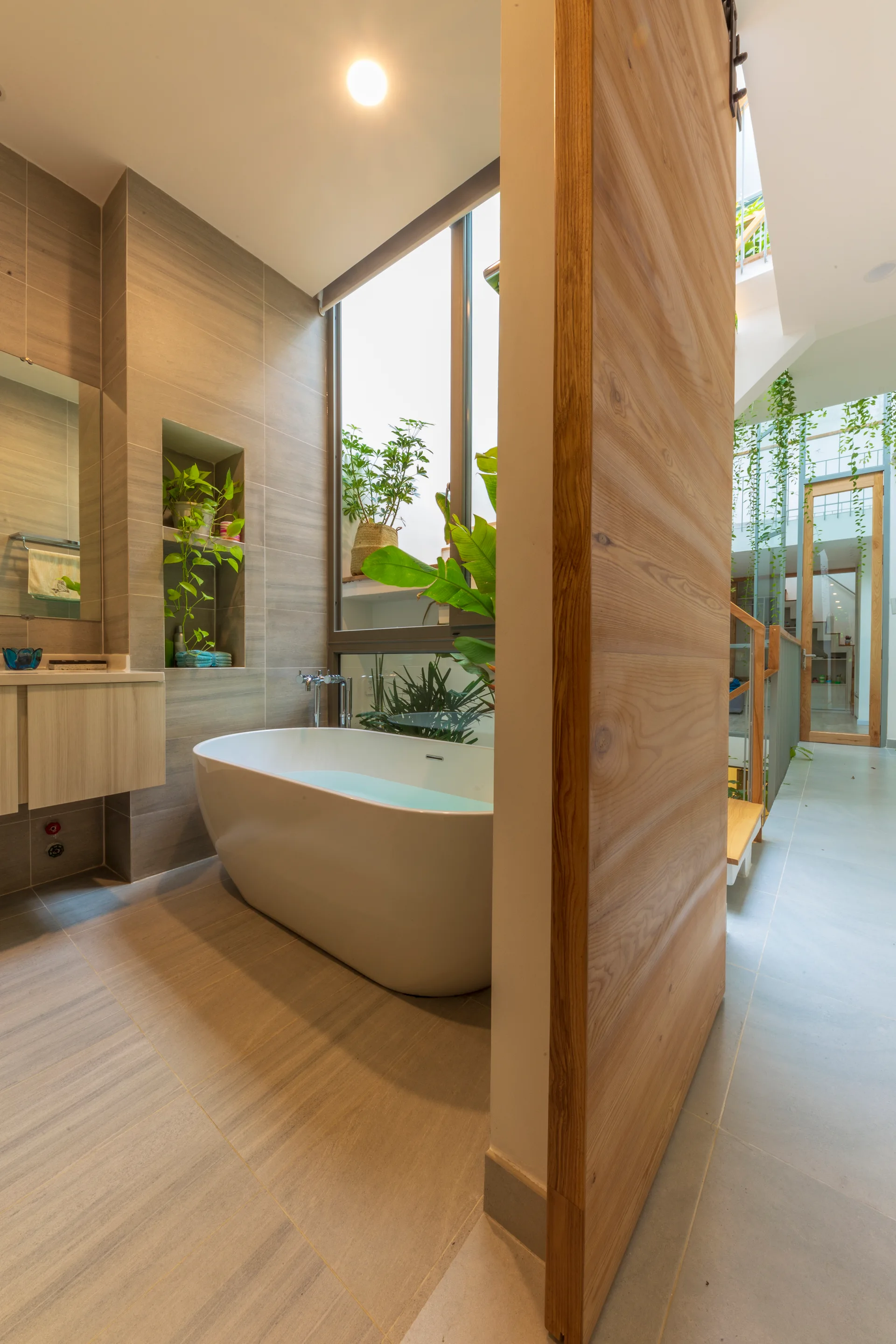A modern bathroom with a freestanding white bathtub, a recessed plant shelf, a tall window overlooking an atrium, and a large wood partition.