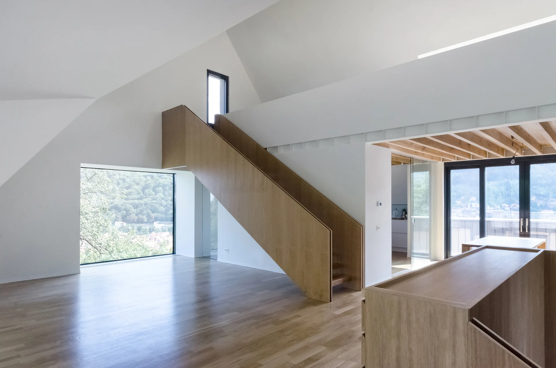 A minimalist living space featuring a custom light wood staircase, vaulted white ceilings, and a large window showcasing a lush forest landscape.