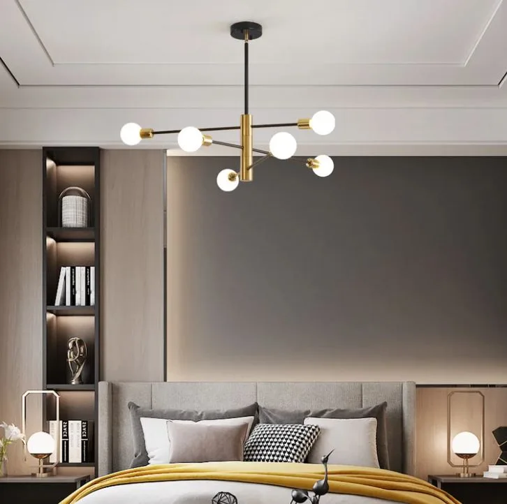 A modern bedroom features a brass globe chandelier hanging over a bed with a grey upholstered headboard and warm cove lighting on the wall panel.