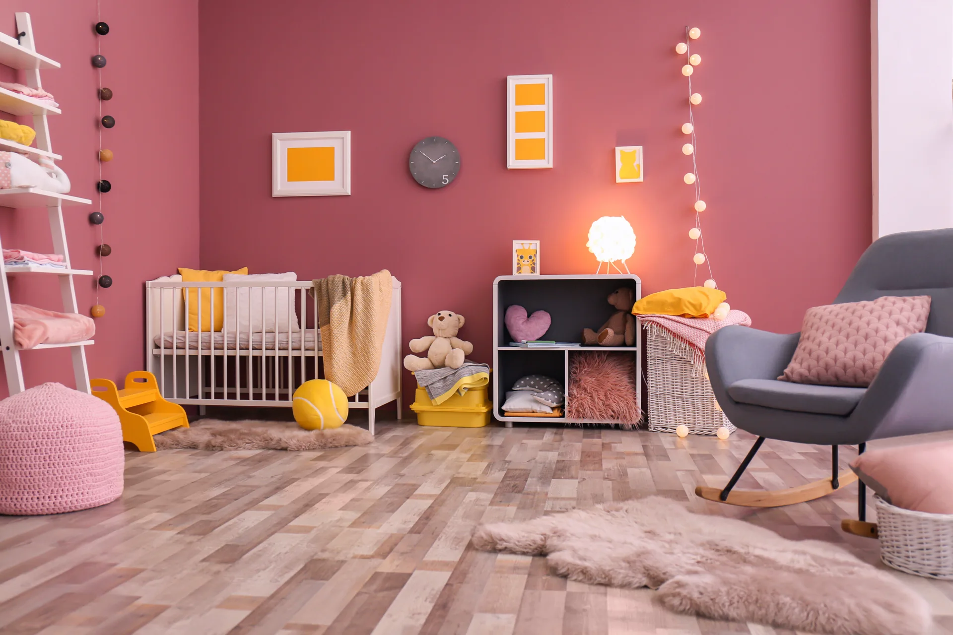 A cozy nursery features a white crib, a grey rocking chair, and open shelving against a mauve wall accented with yellow decor and soft string lights.