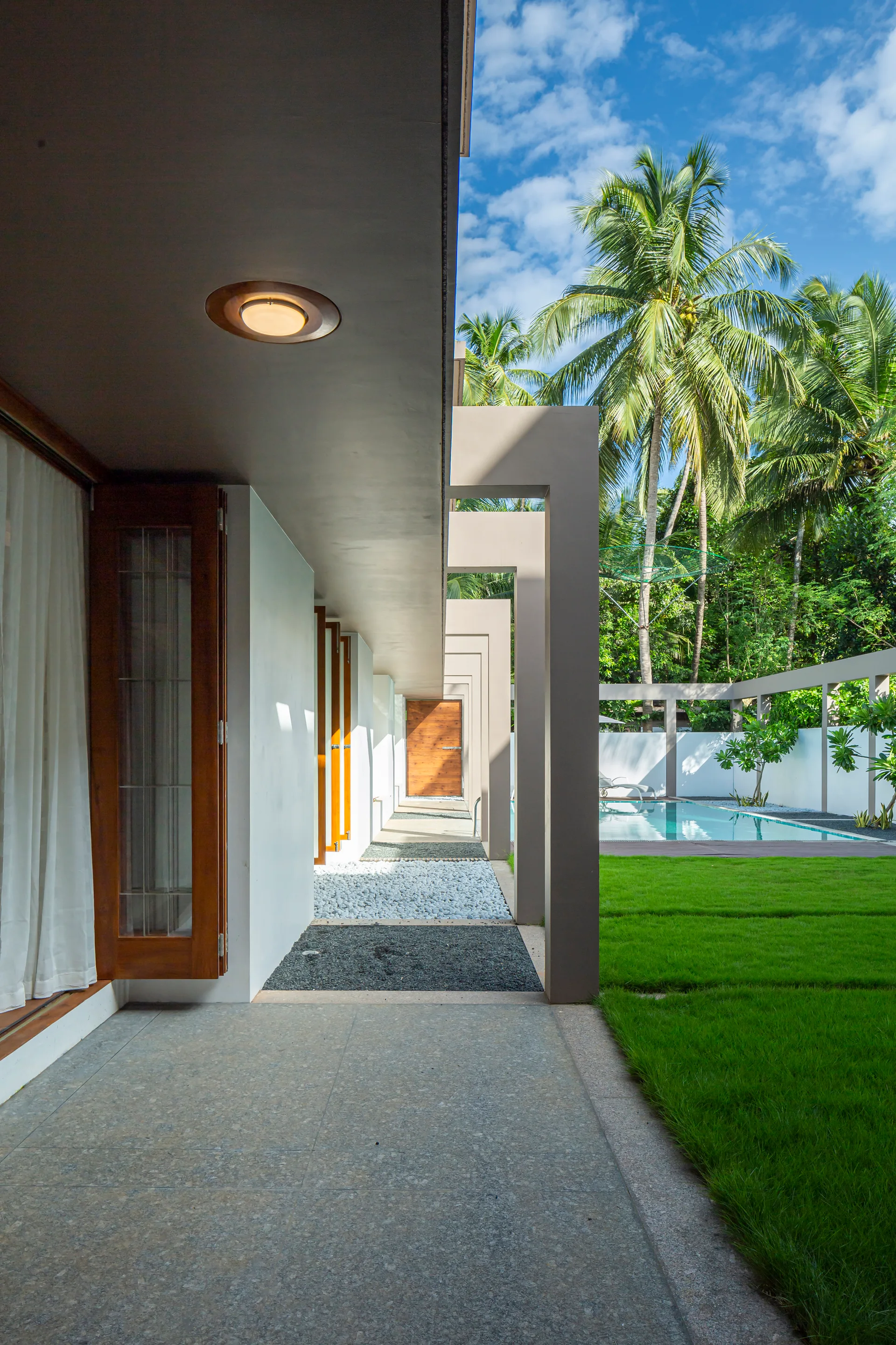 A covered modern concrete walkway with geometric pillars runs alongside a green lawn, pebble landscaping, and a blue swimming pool in a tropical setting.