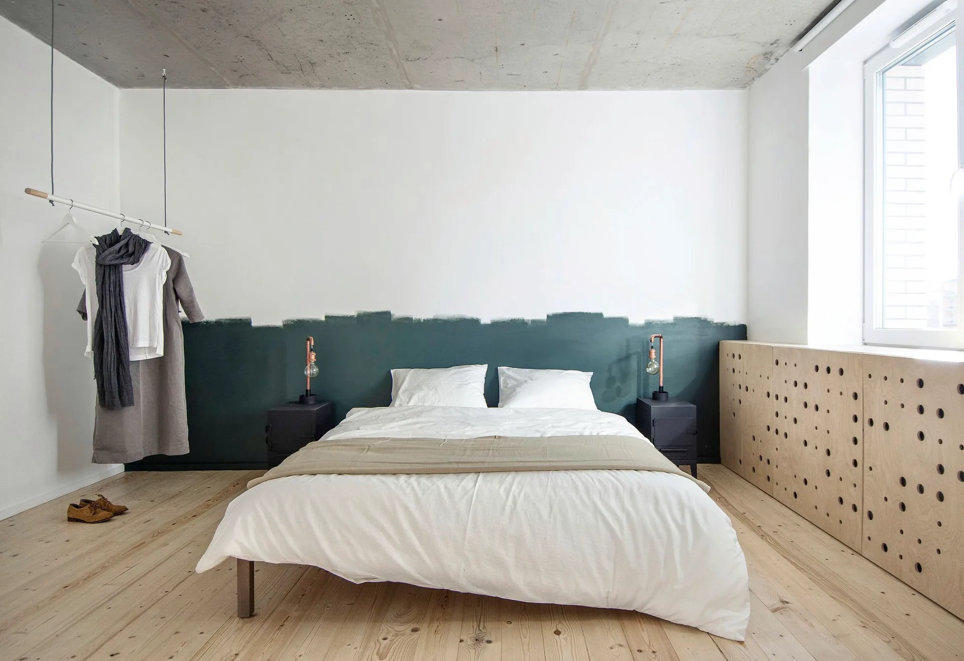 Minimalist bedroom featuring a bed with white linens beneath an exposed concrete ceiling, a dark green painted wall band, and a perforated plywood cabinet.