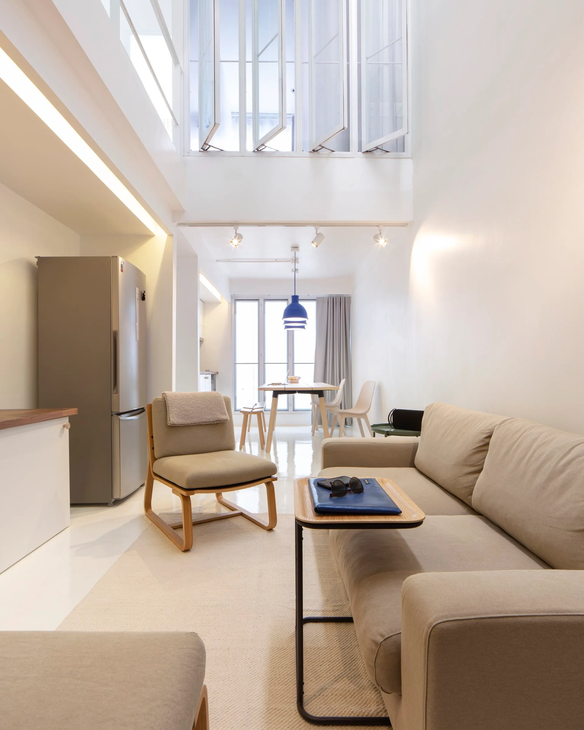 A bright, open-plan minimalist living room features a beige sofa, wood-framed armchair, glossy white floor, and a double-height ceiling.
