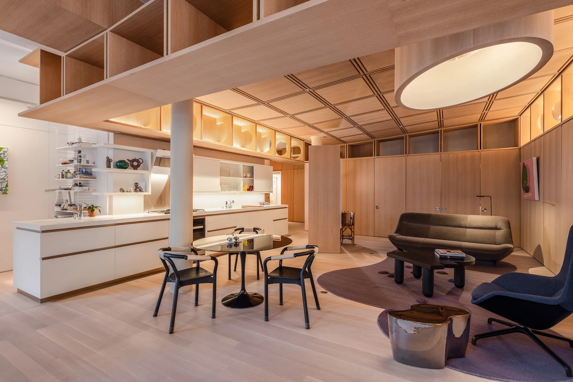 An open-concept kitchen and living area featuring blonde wood wall paneling, a grid-patterned wood ceiling, white cabinetry, and a round dining table.